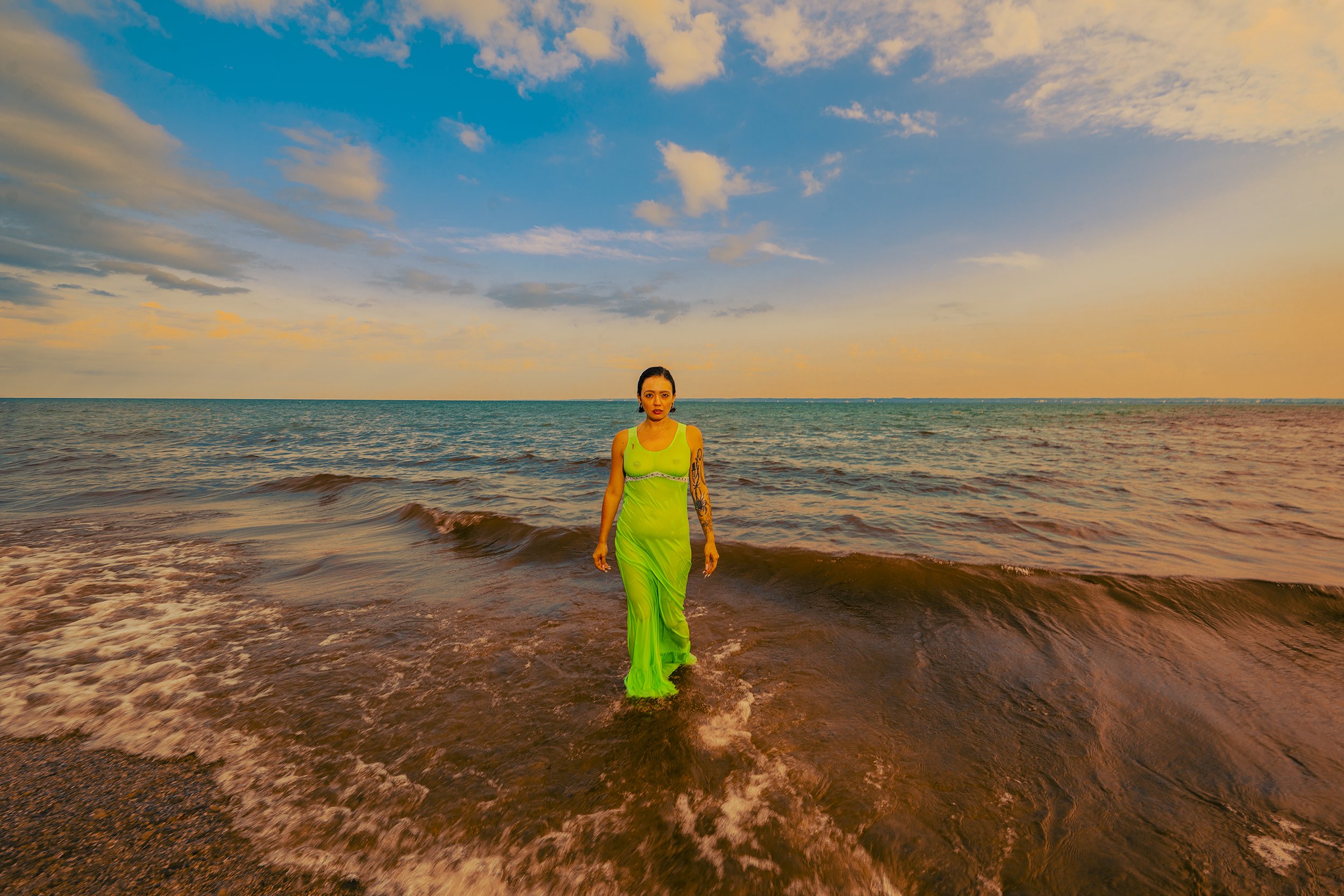 A woman wearing a green dress walking out of the ocean with a sunset sky and clouds above.