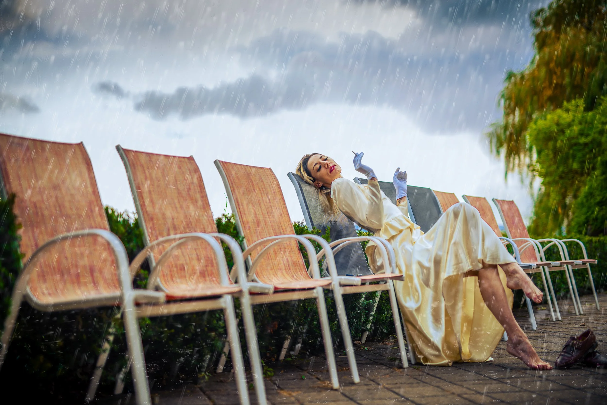 A woman in a yellow dress sitting on chairs outside during a rainstorm, looking relaxed with her eyes closed.