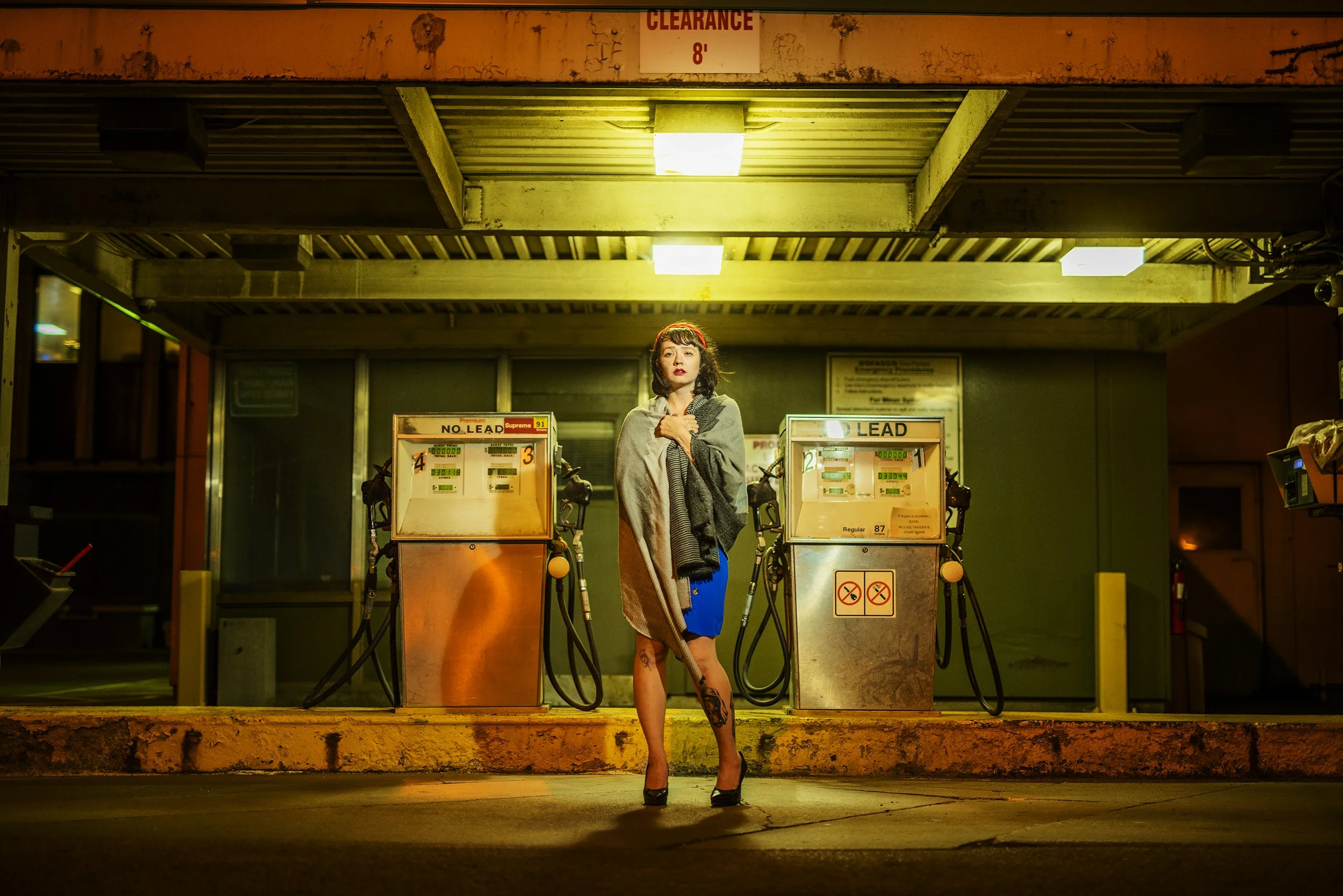A woman with tattoos on her leg stands in front of gas station pumps at night. She is wearing a gray shawl and a blue dress, holding a blanket close to her chest, looking at the camera. The station is lit with yellow lights, and there is a 'Clearance
