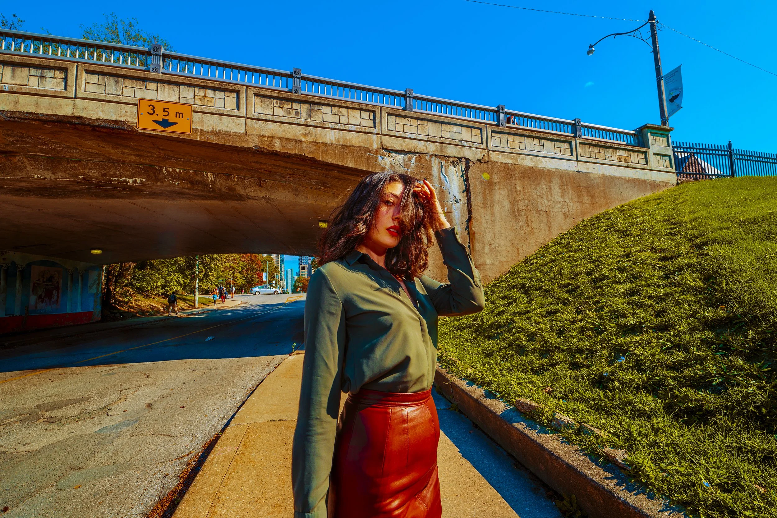 A woman with dark wavy hair wearing a green jacket and red leather skirt standing under a bridge with a blue sky background.