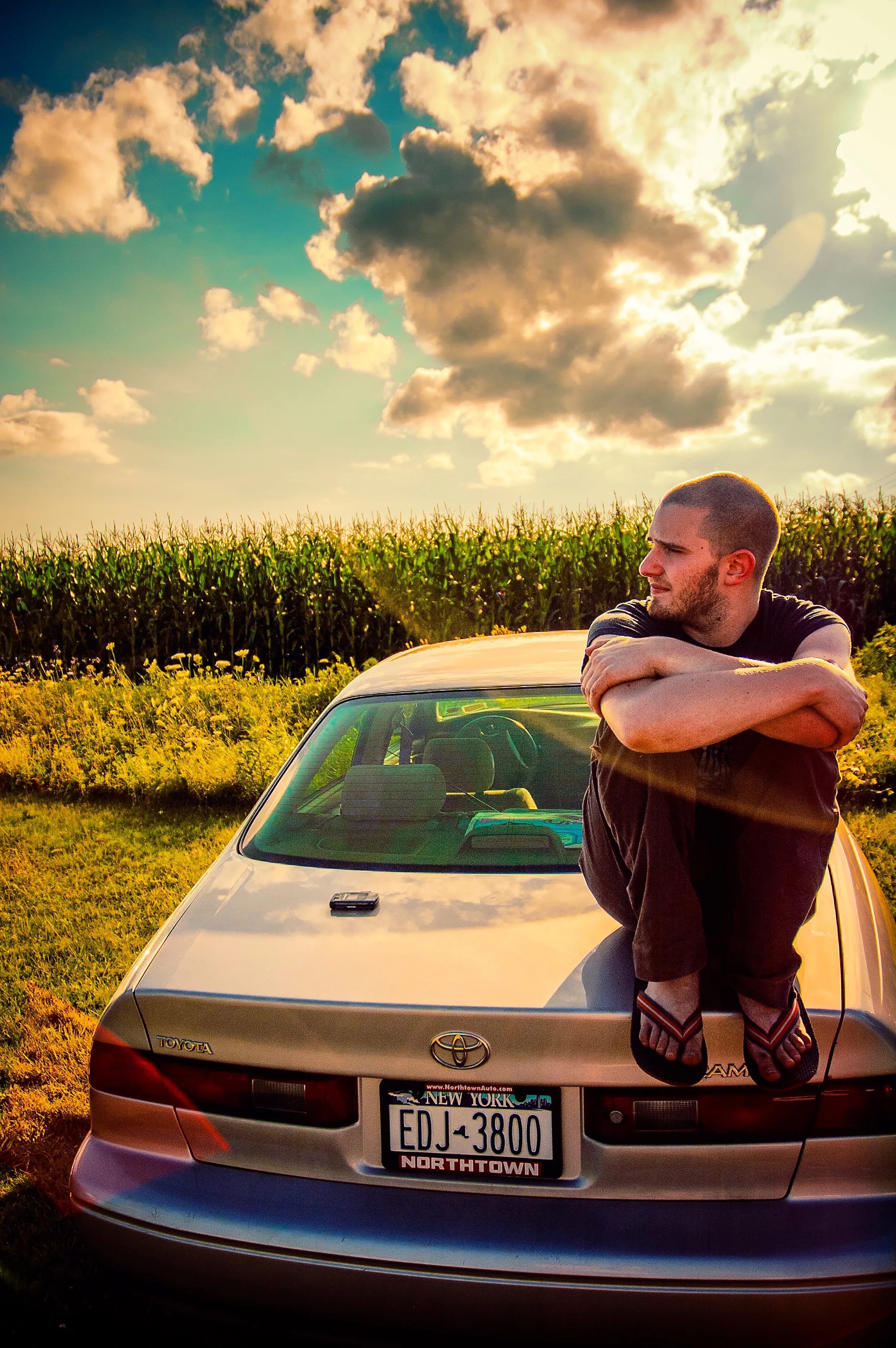 A young man with short hair and a beard is sitting on the hood of a light-colored Toyota sedan with a New York license plate, leaning on his arms, in a field of grass with a cornfield in the background under a sky with dramatic clouds at sunset.