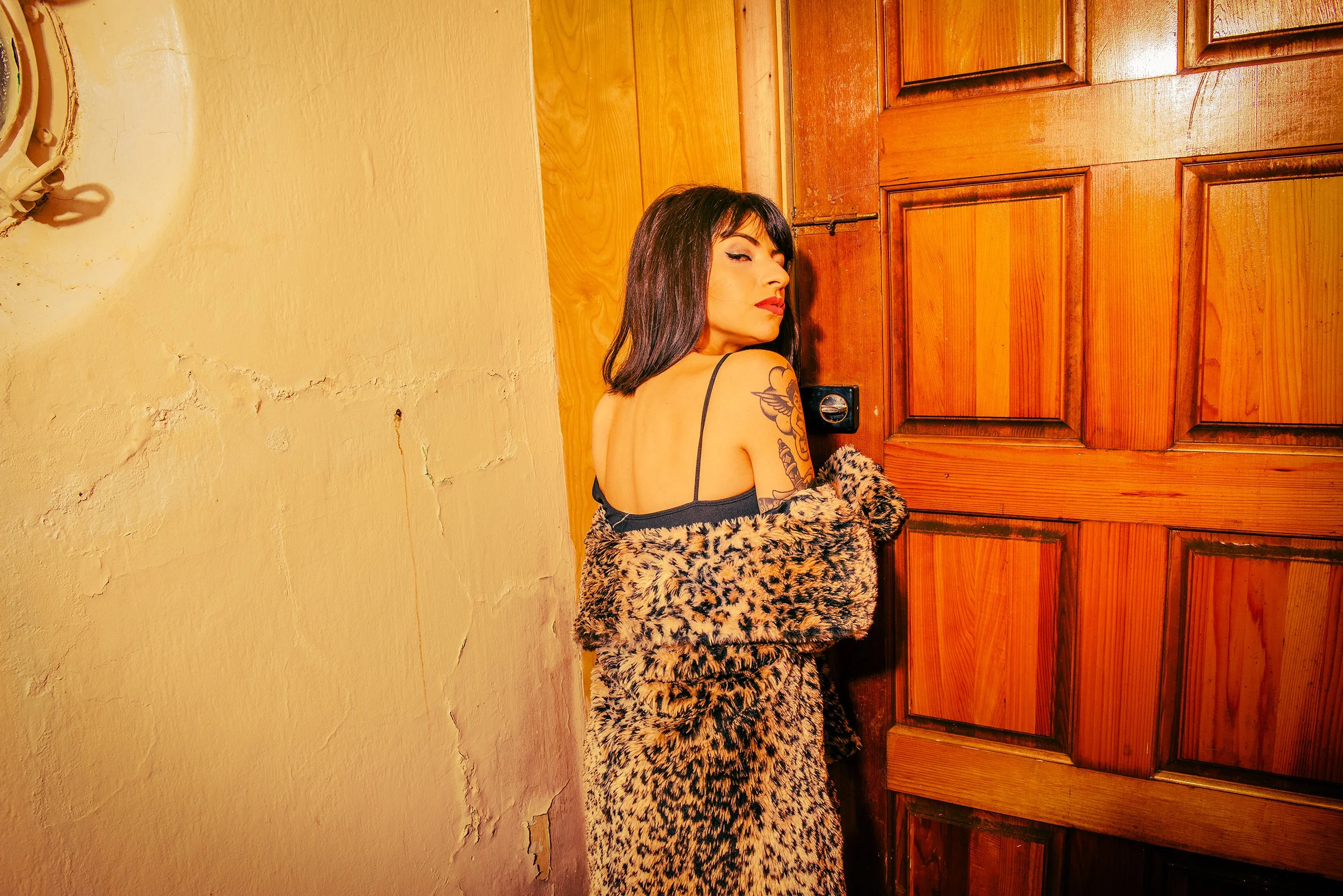 A woman with black hair, tattoos, and lipstick standing against a wooden door, wearing a black spaghetti strap top and a leopard print shawl.
