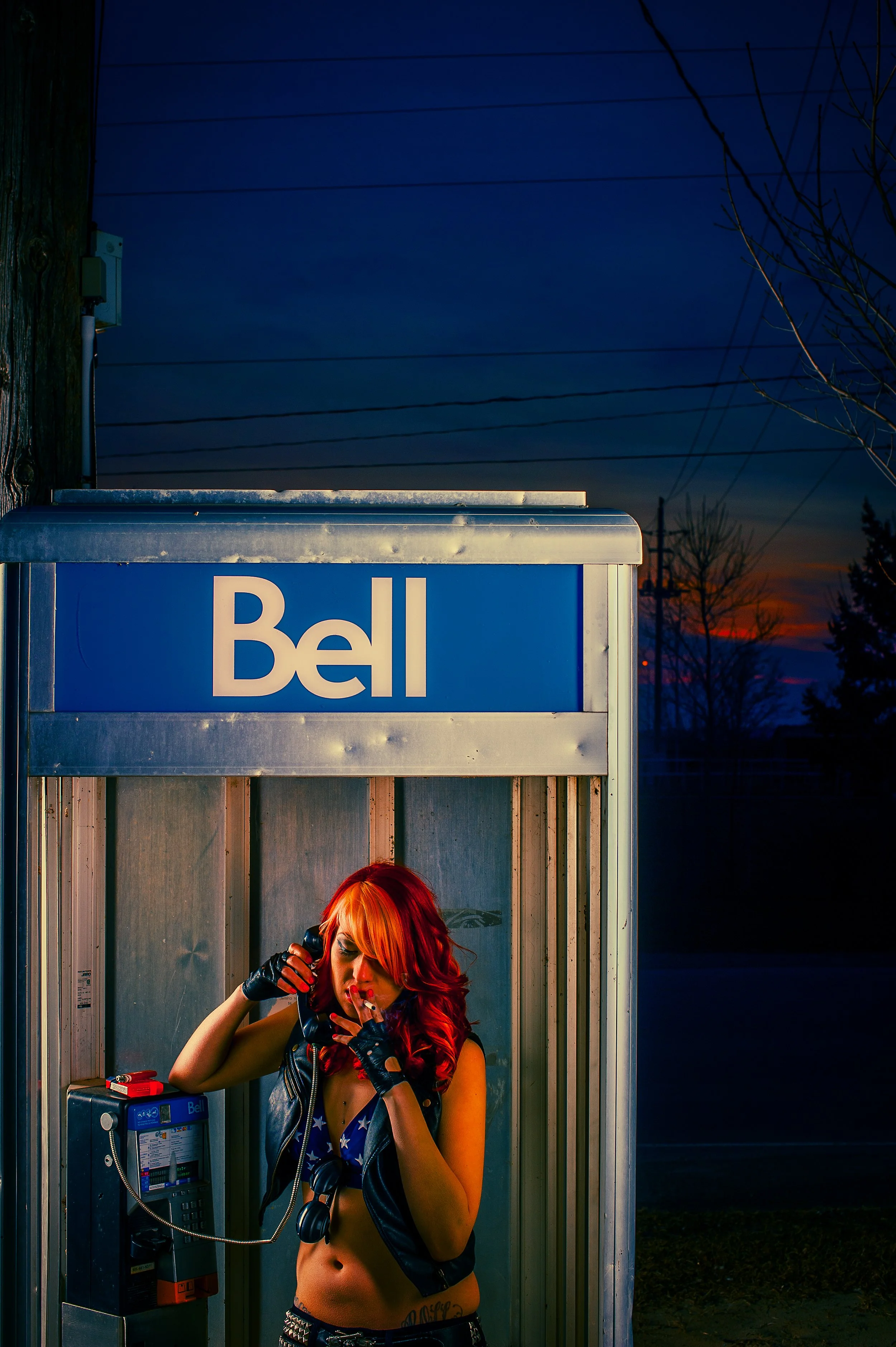 A woman with long red hair talking on a payphone inside a phone booth at dusk. She is wearing a black leather vest with American flag design, fingerless gloves, and has a tattoo on her side. The background shows a darkening sky, power lines, and bare