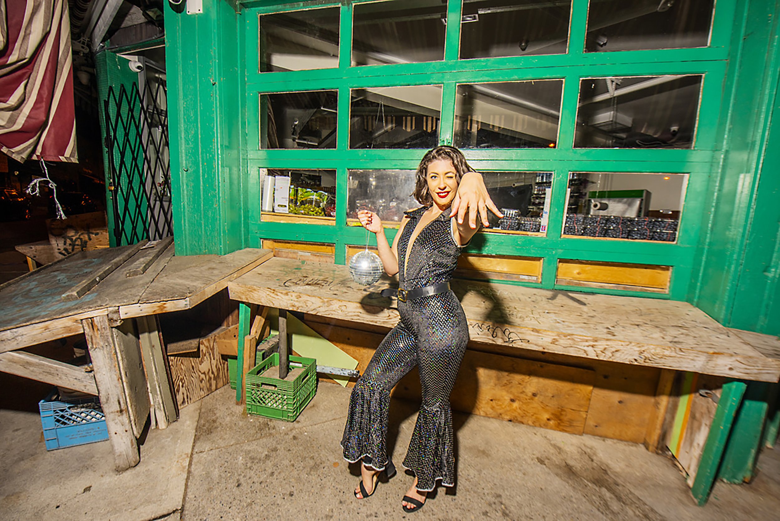 A woman with short curly hair and wearing a sparkly jumpsuit, standing in front of a green-painted wooden structure, reaching out towards the camera and smiling. The setting appears to be outdoors, possibly in an urban area at night, with various cra