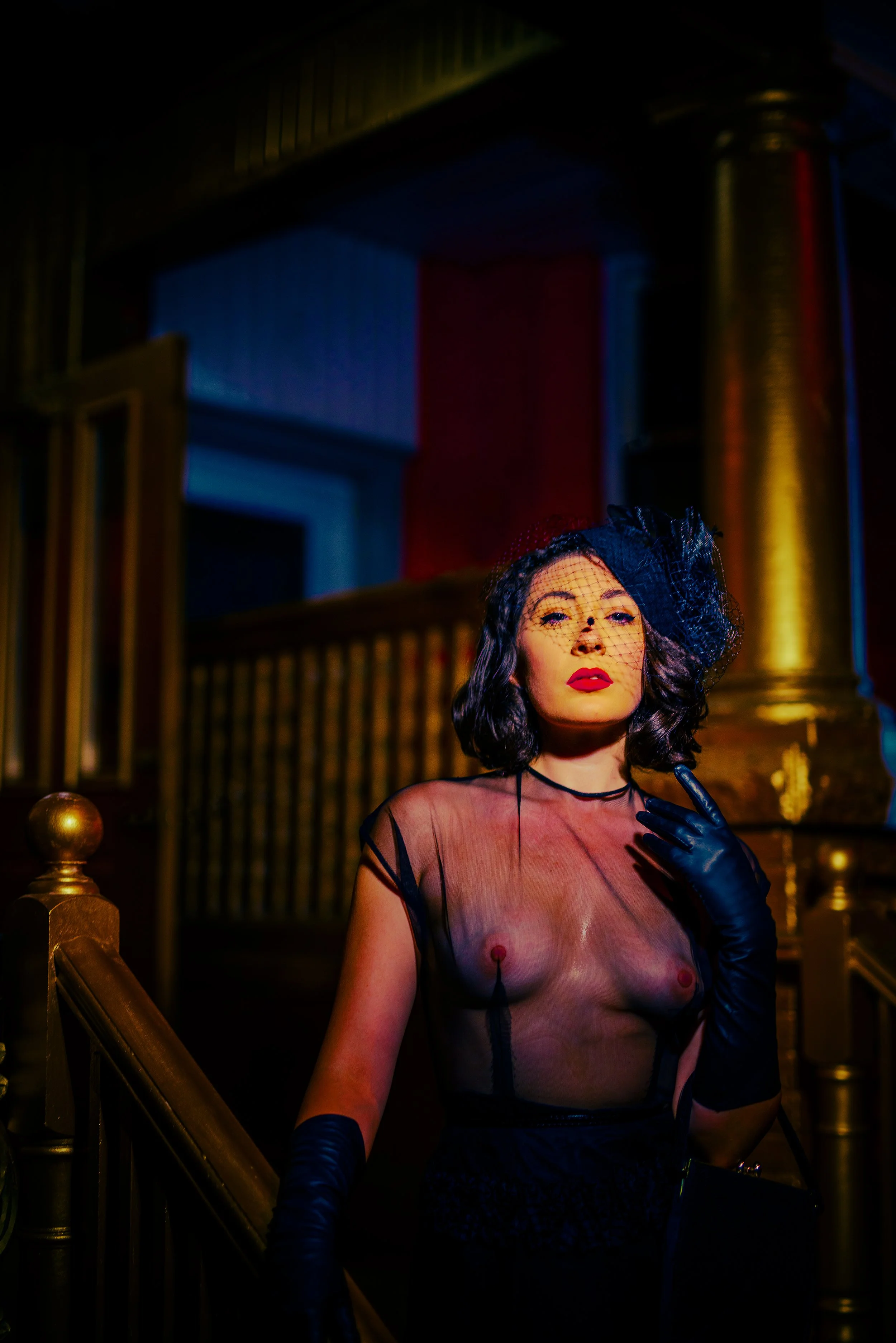 A woman with dark hair and red lipstick, wearing a sheer black top, black gloves, and a black netted fascinator, standing indoors near a staircase with gold accents and playing with her necklace.
