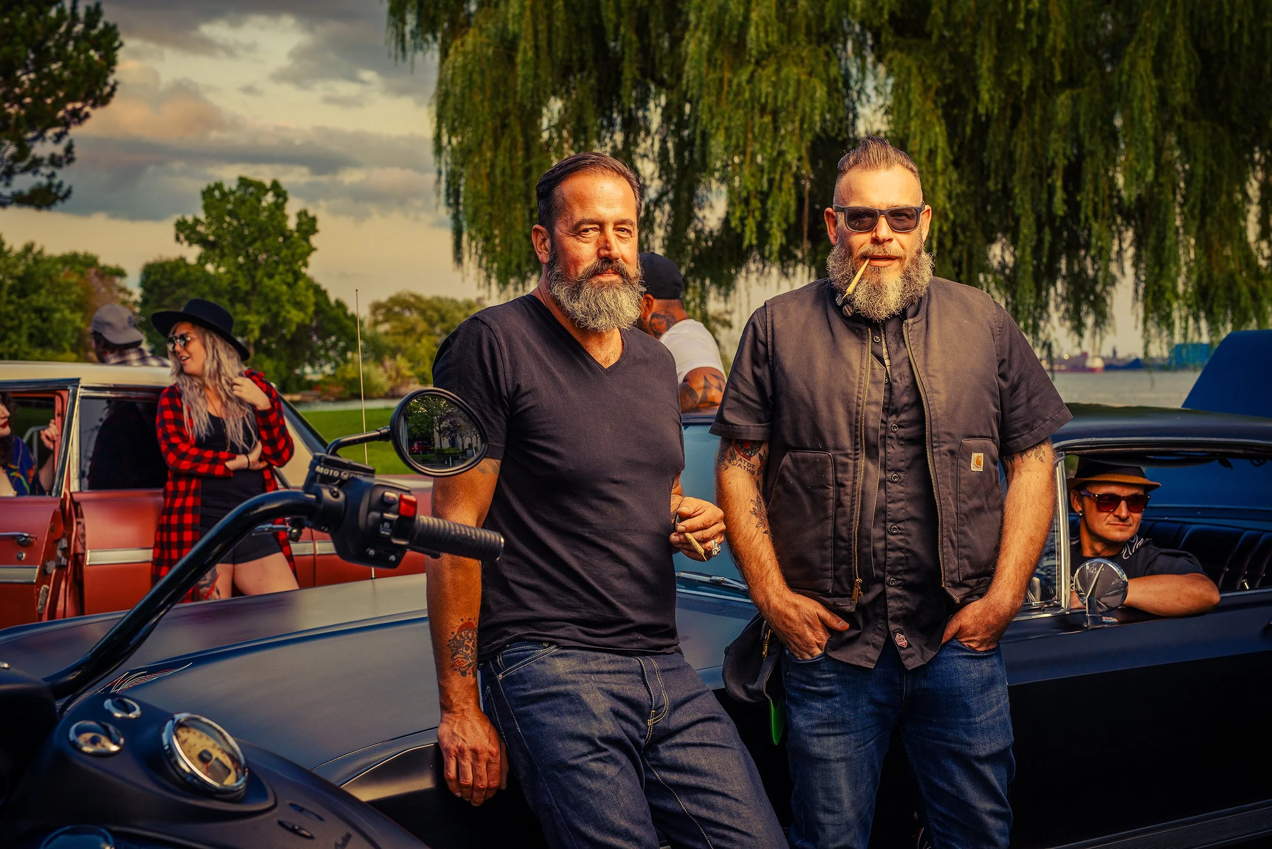 Group of people hanging out outdoors near cars, with two men in the foreground, one with a beard and the other wearing sunglasses and a cigarette in his mouth, under a tree with a lake or river in the background.