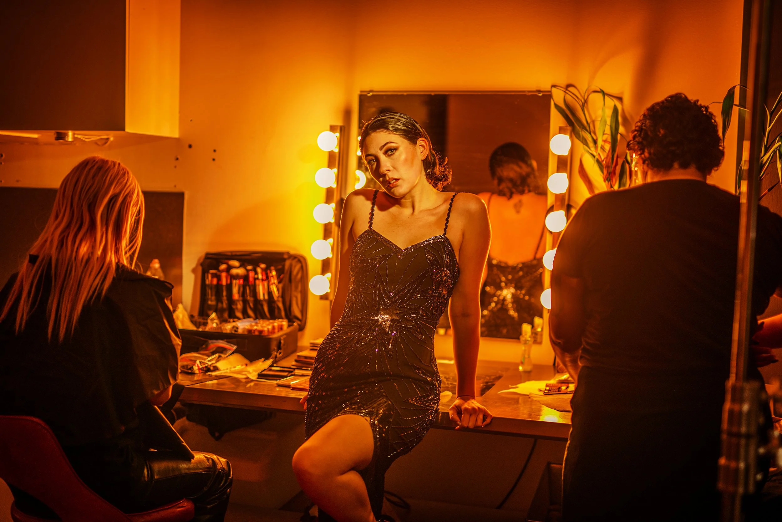 A woman in a black dress poses in front of a mirror with warm orange lighting in a makeup or dressing room, surrounded by two other women.