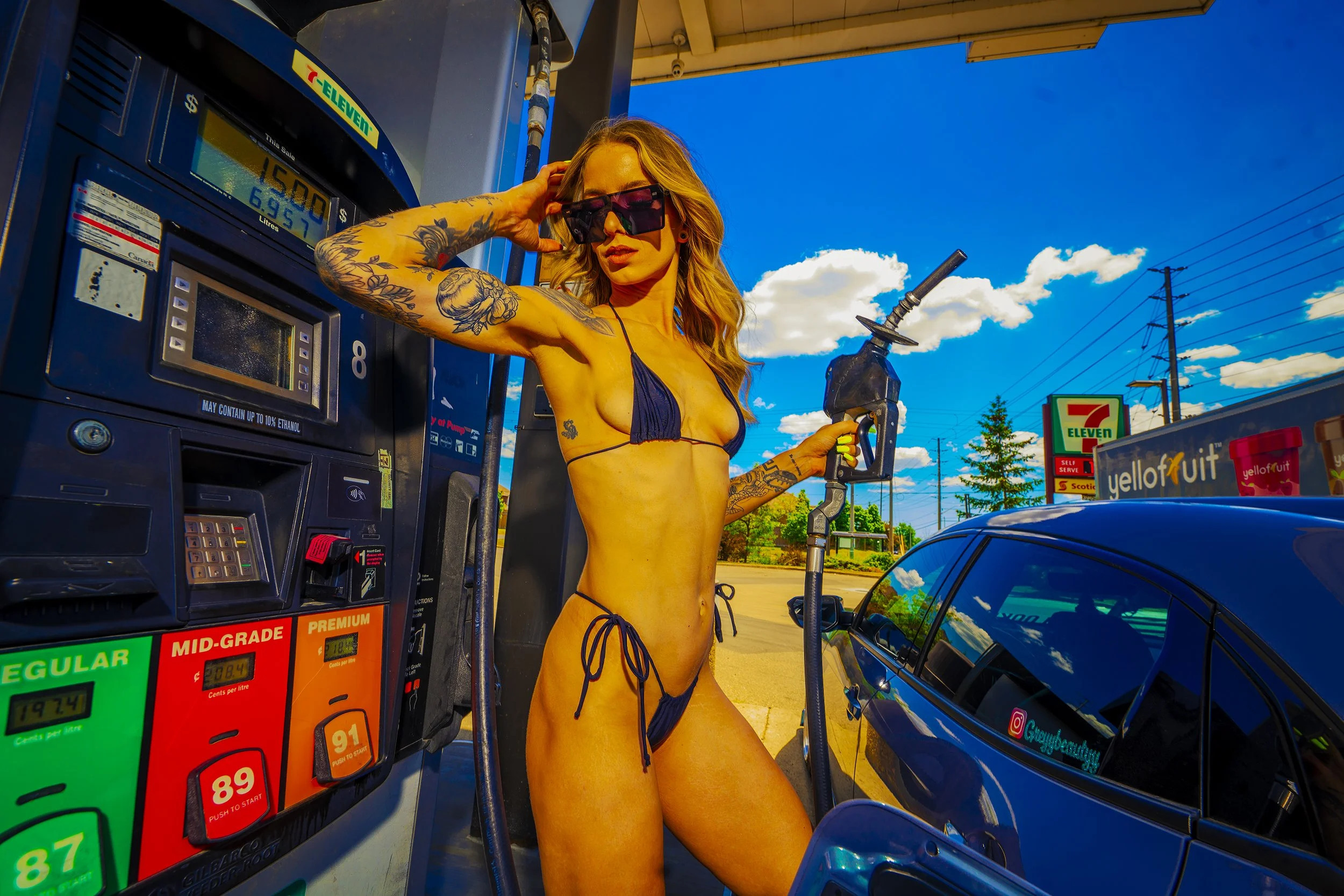 A woman in a blue bikini and sunglasses at a gas station, holding a fuel pump nozzle, with tattoos on her arms, standing next to a black car on a bright sunny day.