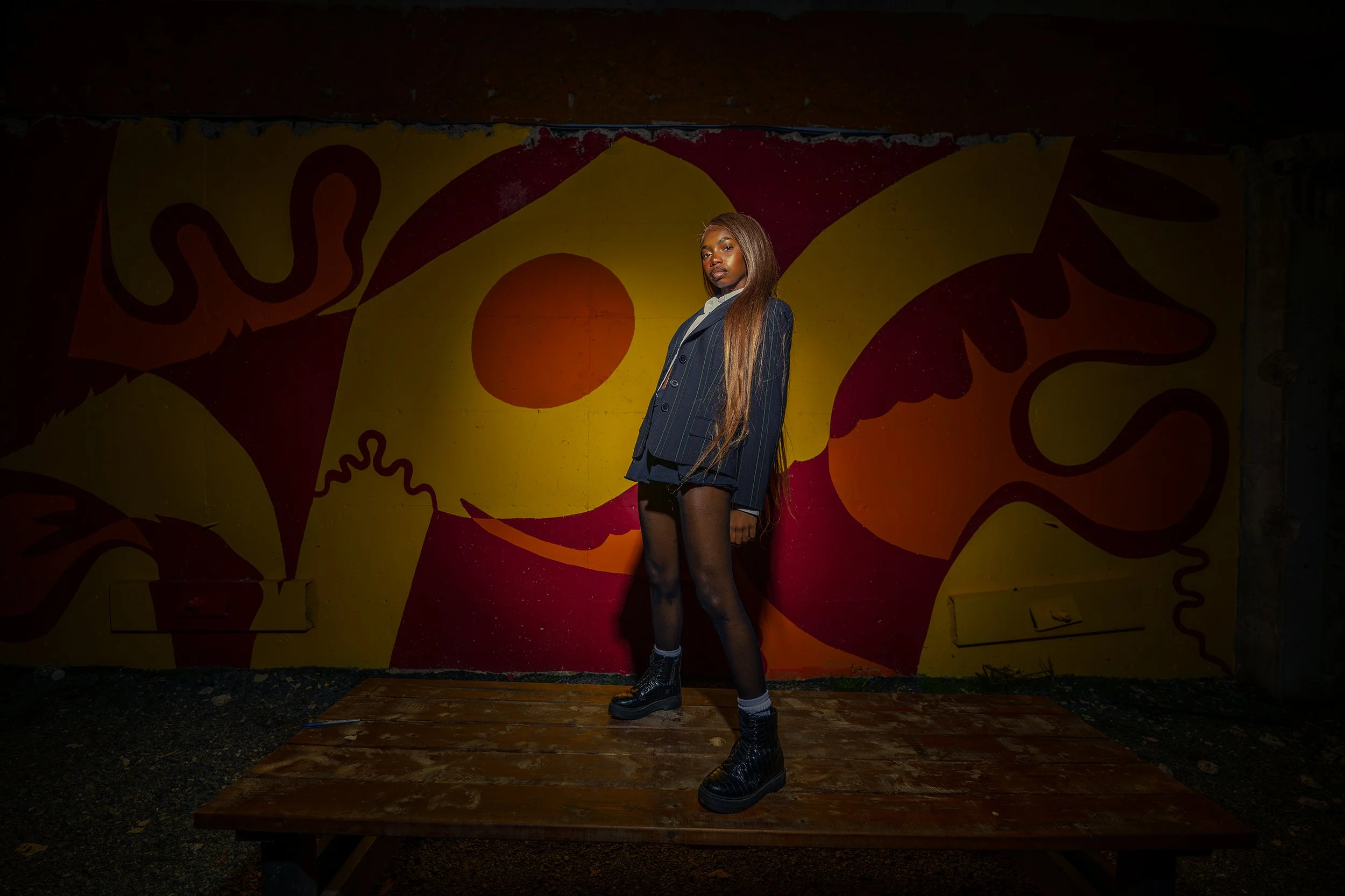 A young woman standing on a wooden platform in front of a colorful abstract mural, wearing a pinstripe blazer, shorts, and combat boots, with long straight hair and a confident expression.