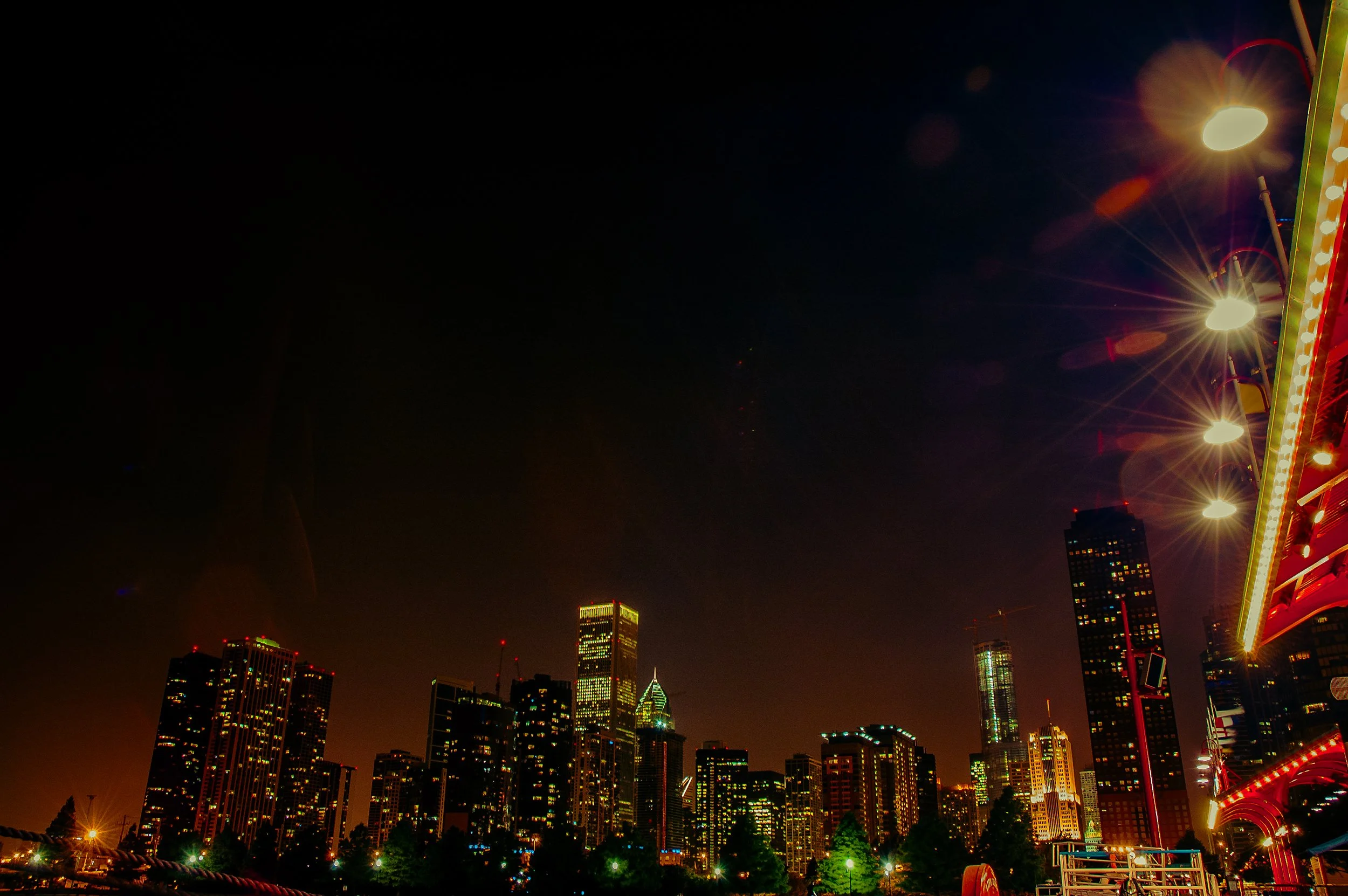 Nighttime city skyline with tall buildings illuminated, a Ferris wheel with bright lights visible on the right, and a dark sky with a few scattered spots of light.