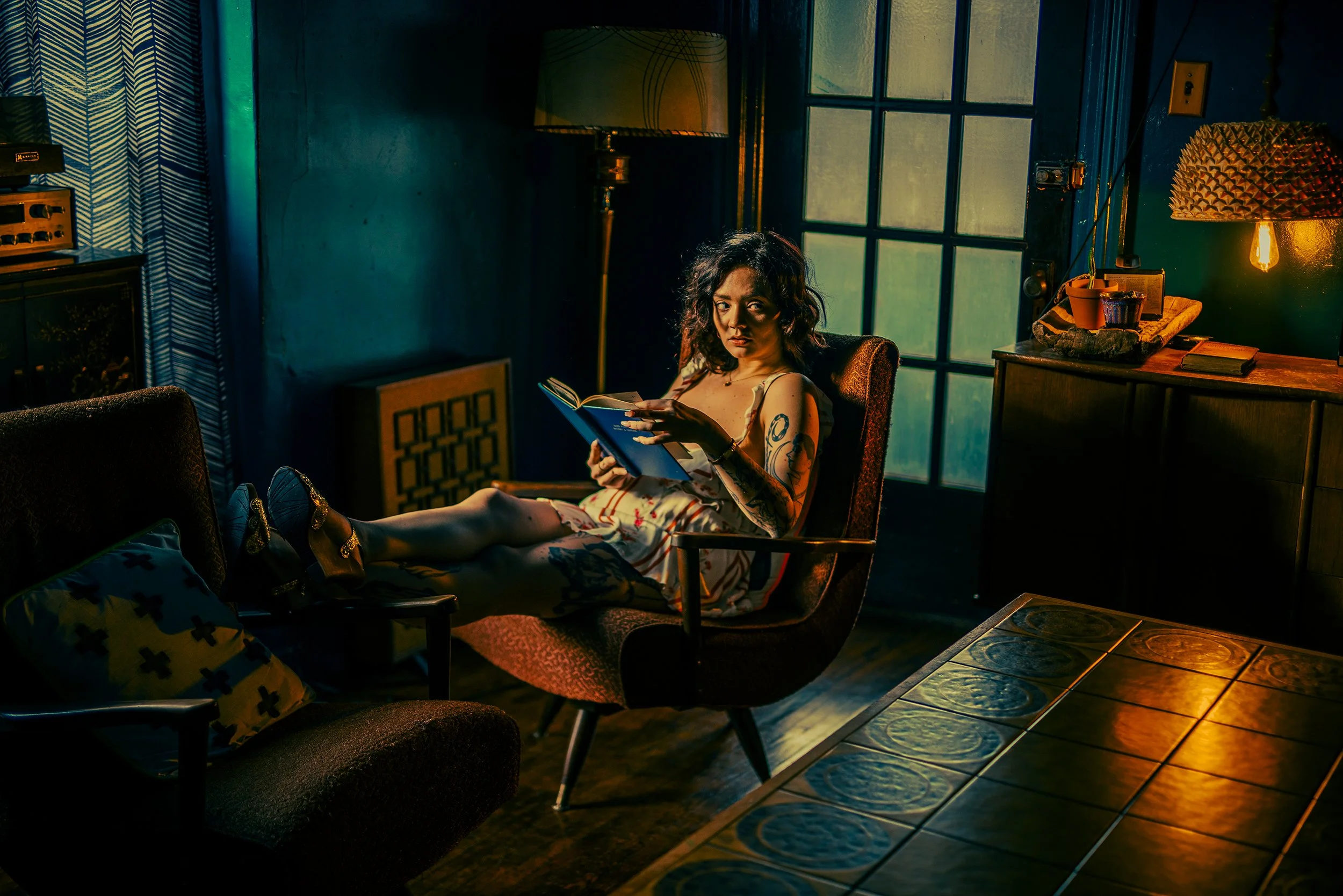 A woman with tattoos and curly hair sitting in a vintage armchair, holding a book, in a dimly lit room with warm lighting, a window, and eclectic decor.