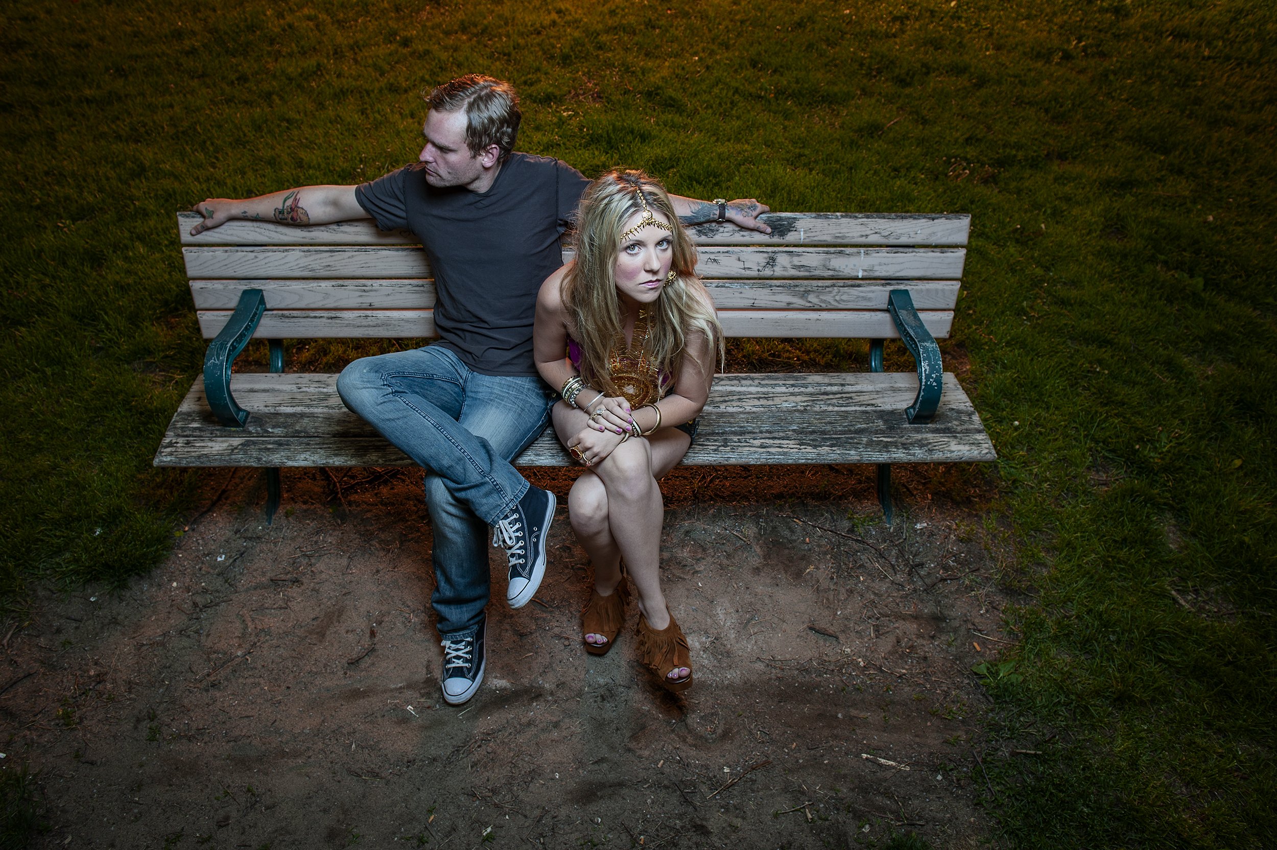 A young couple sitting on a weathered wooden park bench at night. The man has short hair, tattoos on his arm, and is wearing a black T-shirt and jeans. He is sitting sideways on the bench, looking at the woman. The woman has long blonde hair, wearing