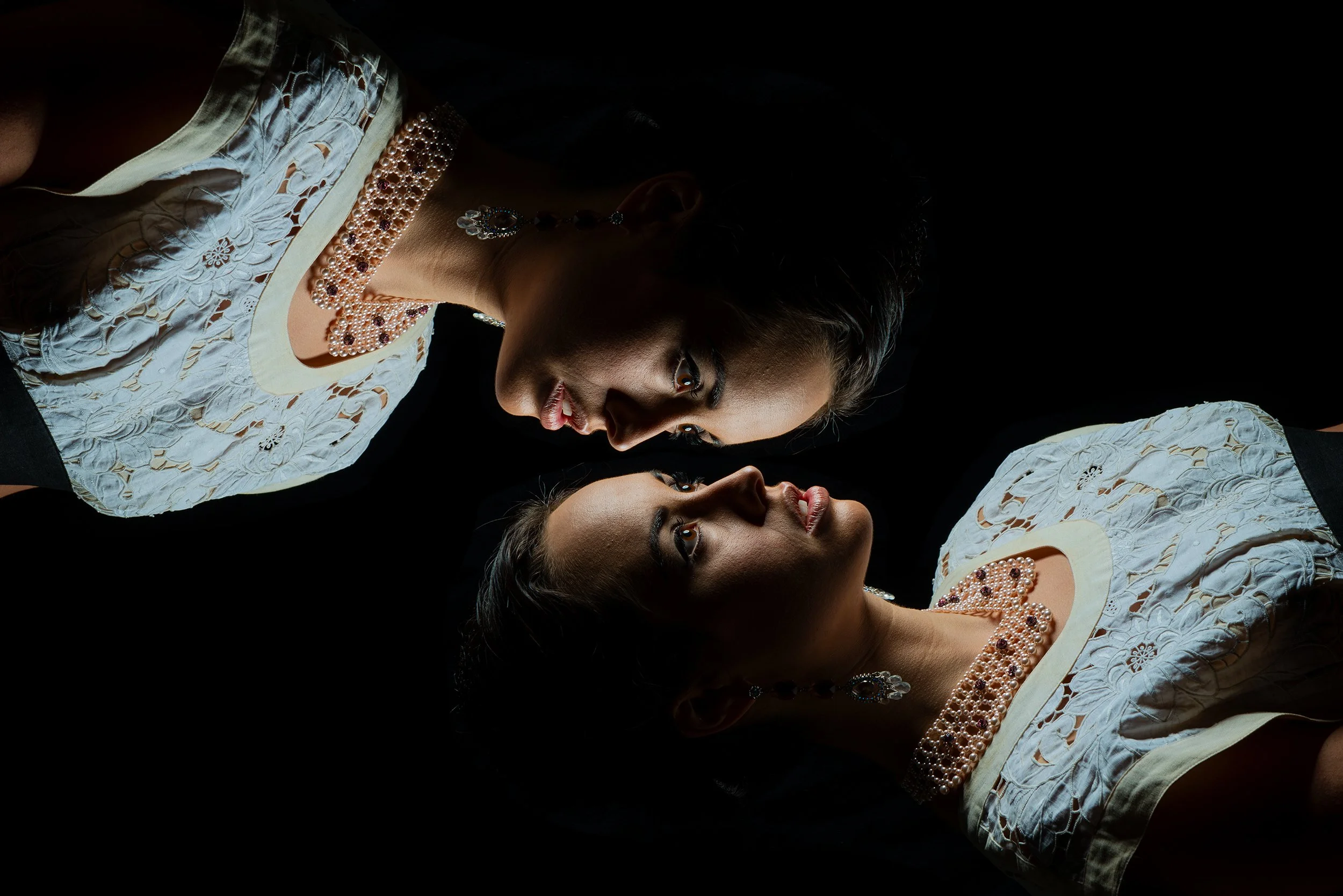 The image shows two women with dark hair and brown skin, dressed in traditional white embroidered outfits, facing each other with their heads tilted toward each other. They are wearing pearl and gemstone jewelry, including necklaces and earrings, wit