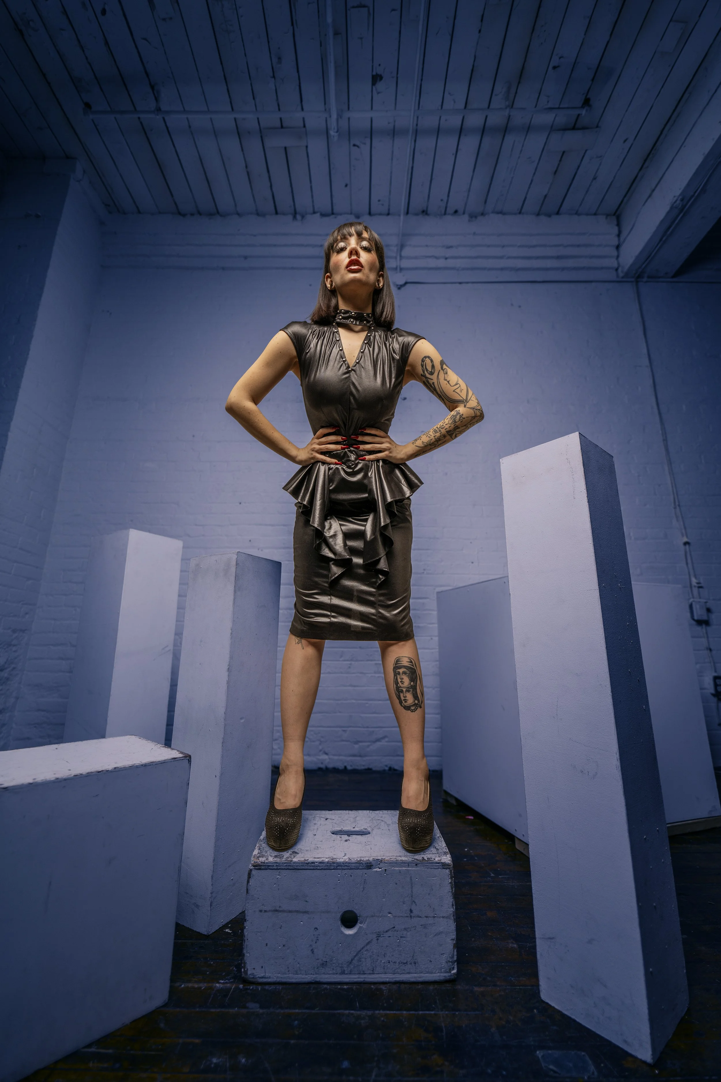 A woman in a black leather dress and high heels standing on a platform with her hands on her hips in an industrial-looking room with white walls and white pedestals.
