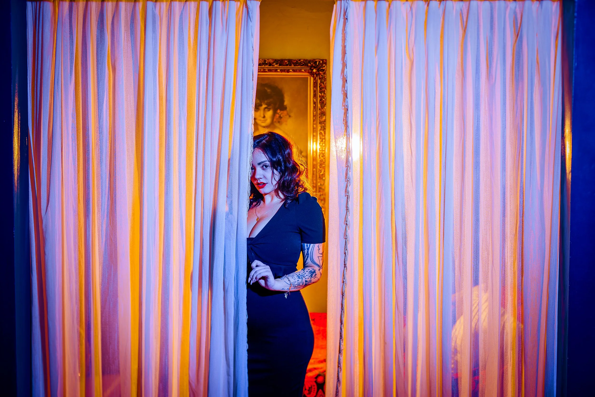 A woman with dark wavy hair and tattoos on her arm, wearing a black top, is partially hidden behind striped curtains, with a framed portrait of a woman with curly hair in the background. The scene is illuminated with warm, colorful lighting.