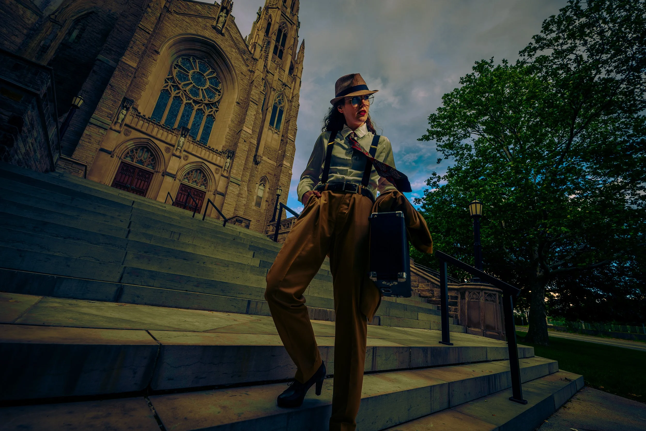 A woman in vintage clothing, wearing a hat, sunglasses, and suspenders, standing on the steps outside a historic church with Gothic architecture and stained glass windows.