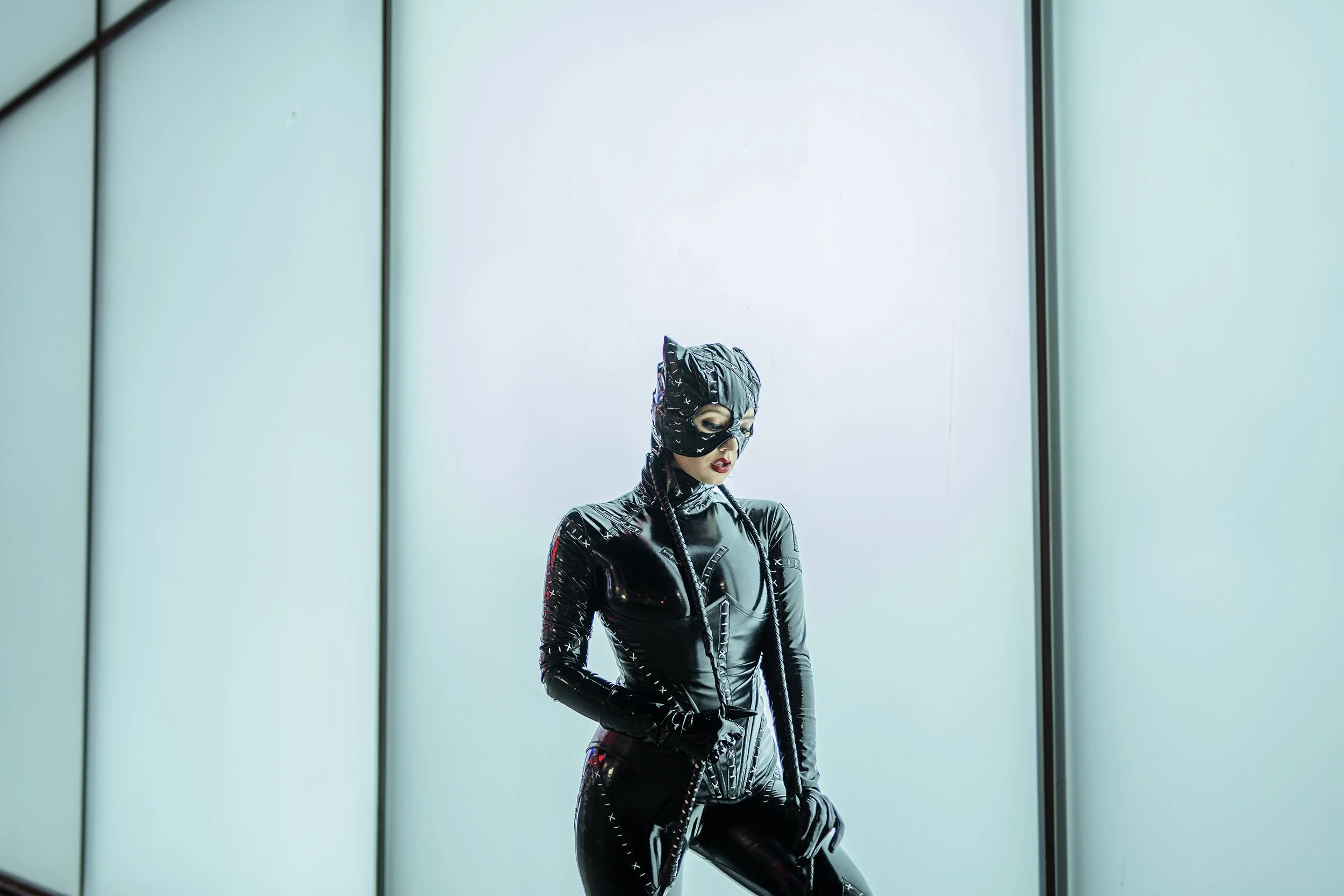 A woman dressed in black, vinyl-like outfit with a matching mask, standing in an elevator with teal-tinted glass panels.