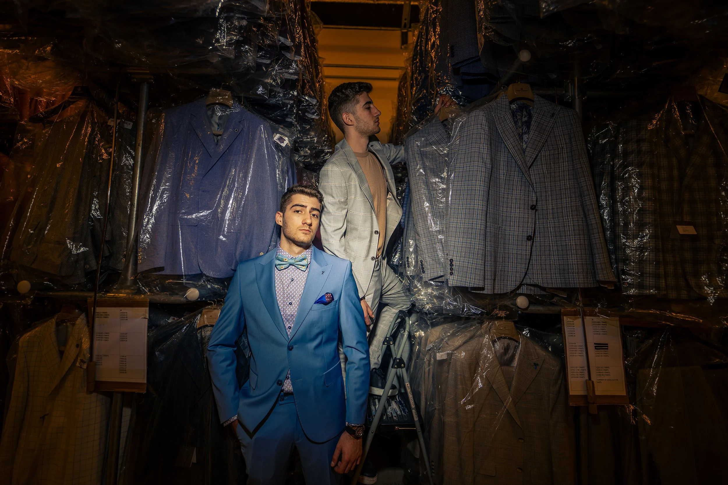 Two young men shopping for suits in a clothing store, surrounded by racks of plastic-covered jackets and blazers, with one man looking at a coat on a hanger and the other standing in front wearing a bright blue suit.