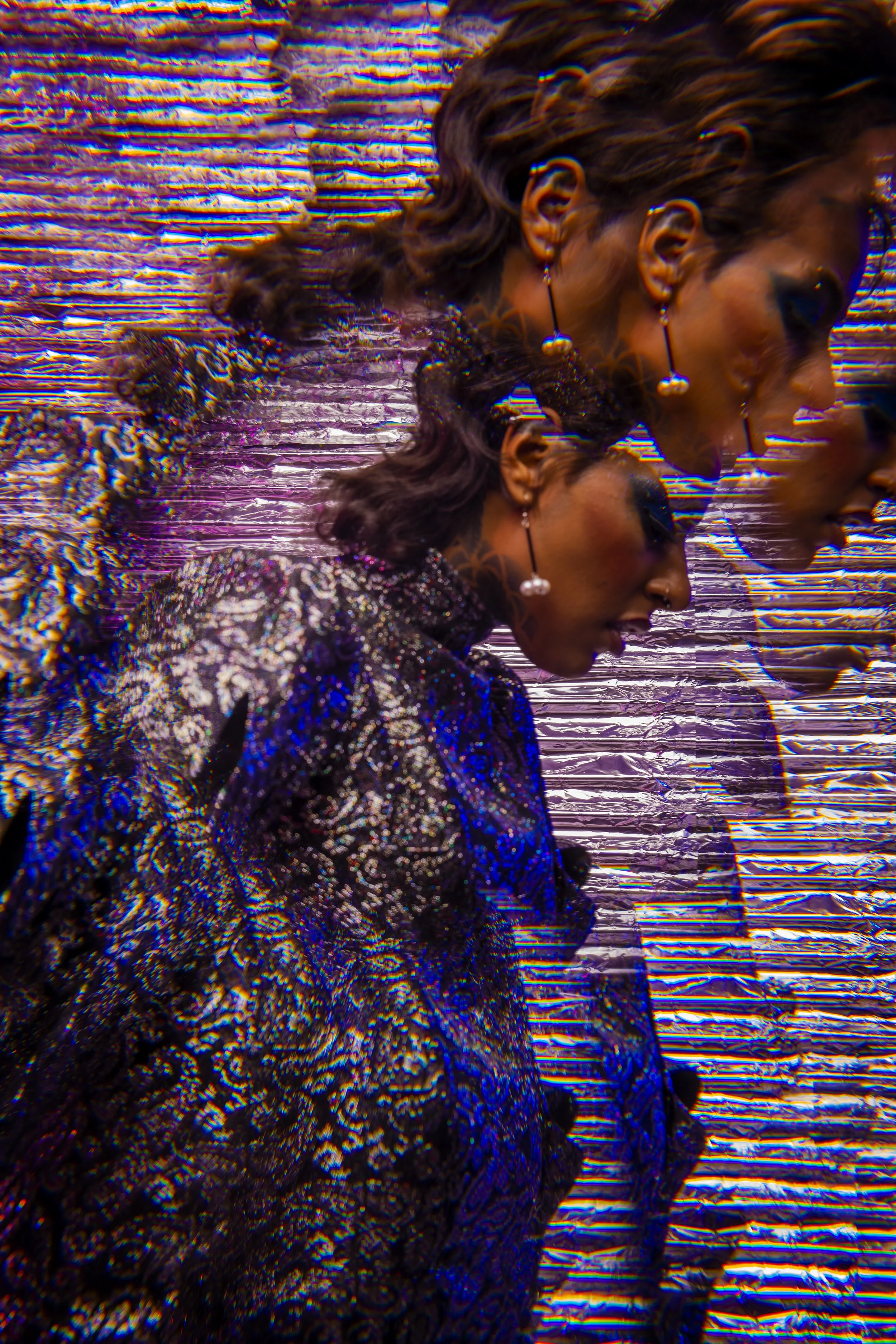 Two women dressed in sparkly clothing and wearing earrings, with distorted colorful digital effects, facing to the right against a shiny, metallic background.