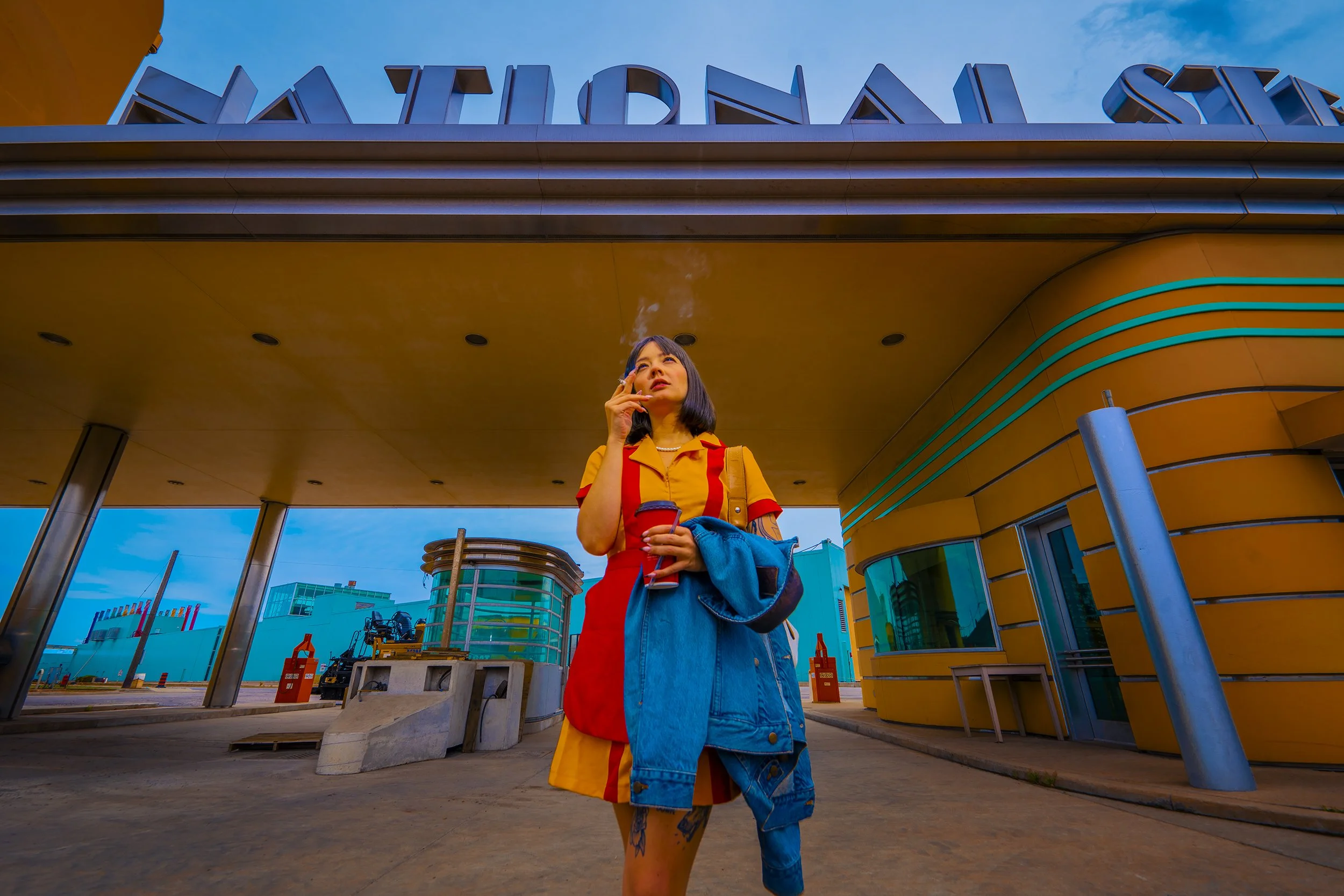 A woman in a colorful outfit standing under a modern building with the word 'FAIRVIEW' on top, smoking a cigarette, holding a drink, and carrying a denim jacket.