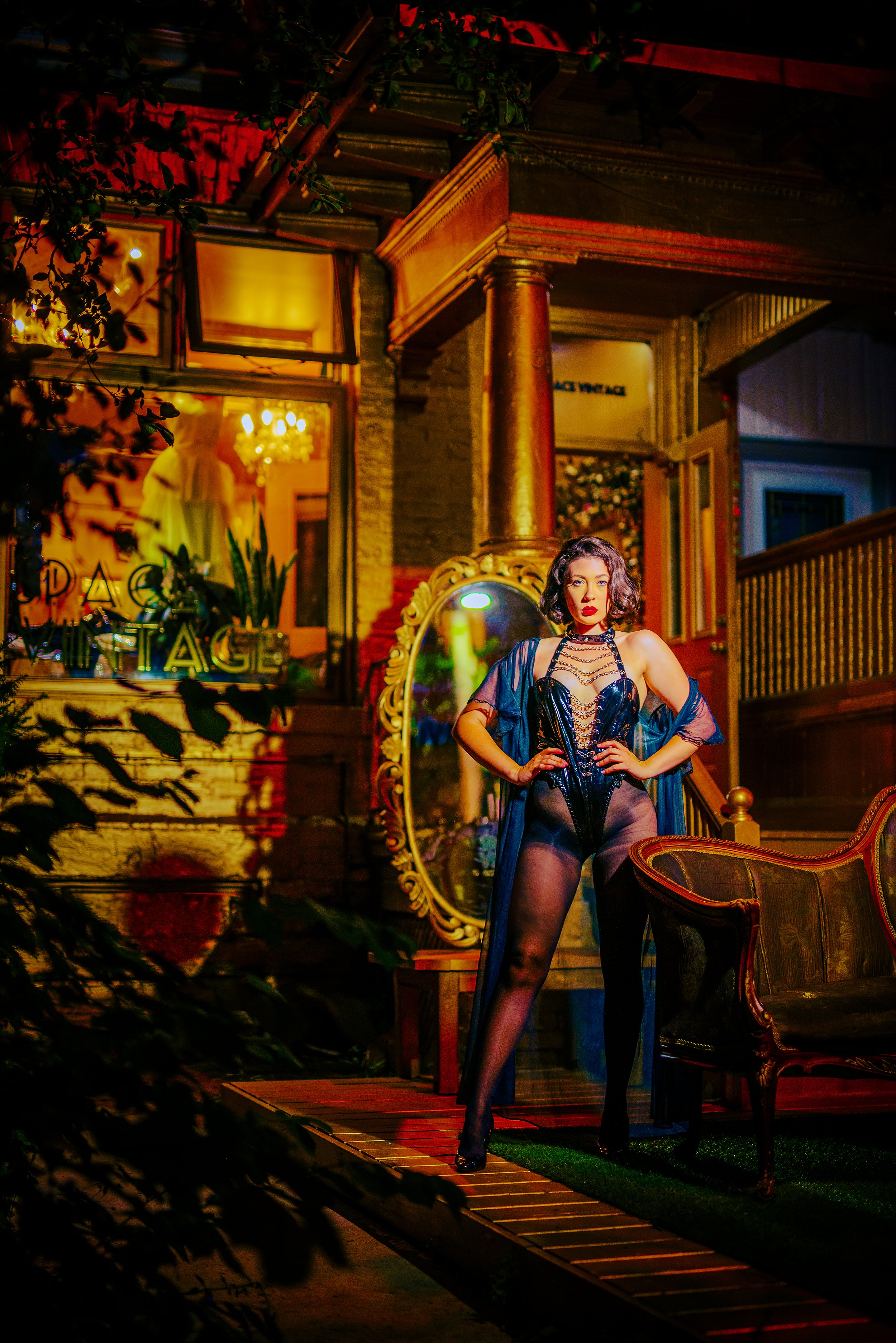 A woman with dark, curled hair and bold makeup, wearing a black leather corset, lace stockings, and a sheer dark robe, standing confidently in a vintage-style room illuminated with warm, colorful lighting. The room features antique furniture, mirrors
