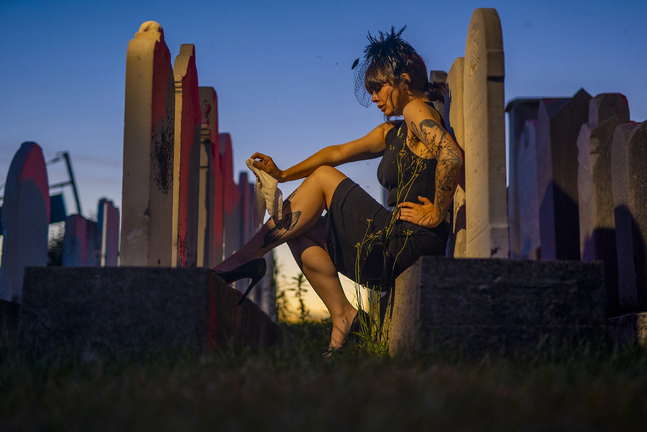 A woman with tattoos in a black dress and high heels sitting on a grave at sunset, holding a white cloth with tombstones and a blue sky in the background.