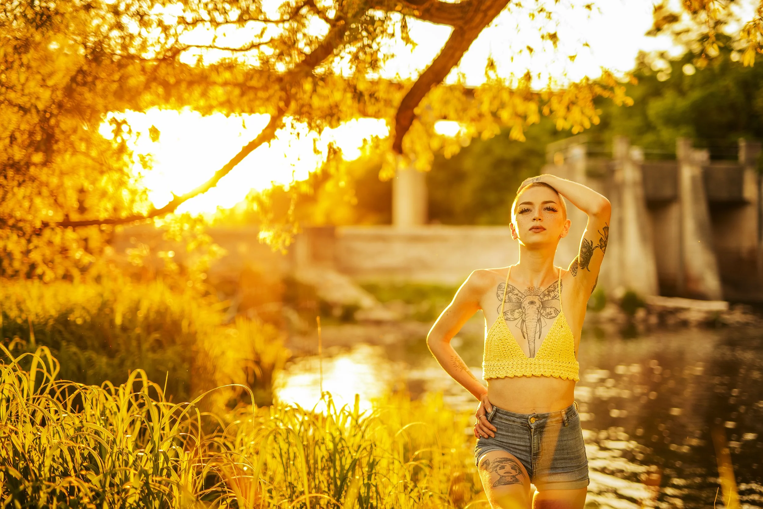A woman with tattoos wearing a yellow crochet top and denim shorts standing outdoors near a river during sunset, with trees and a bridge in the background.