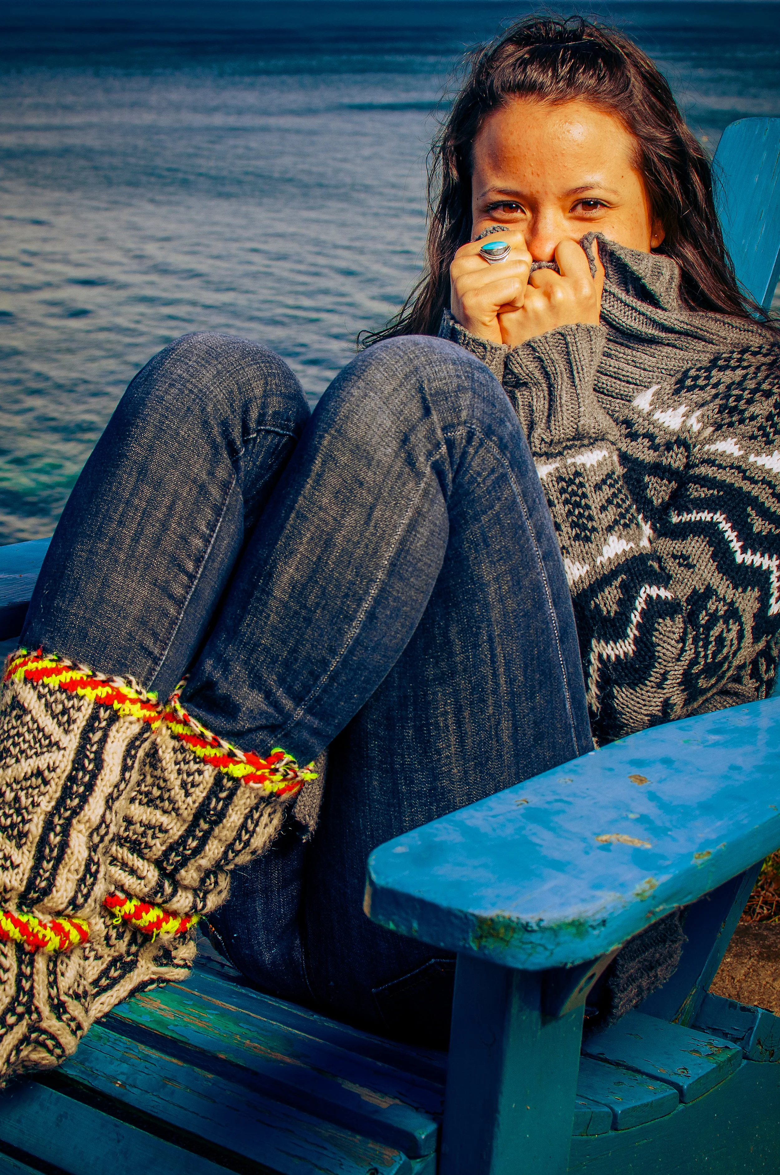 A young woman with dark hair and freckles sitting on a blue wooden bench near water, wearing a patterned sweater and pajamas, smiling with tears in her eyes.