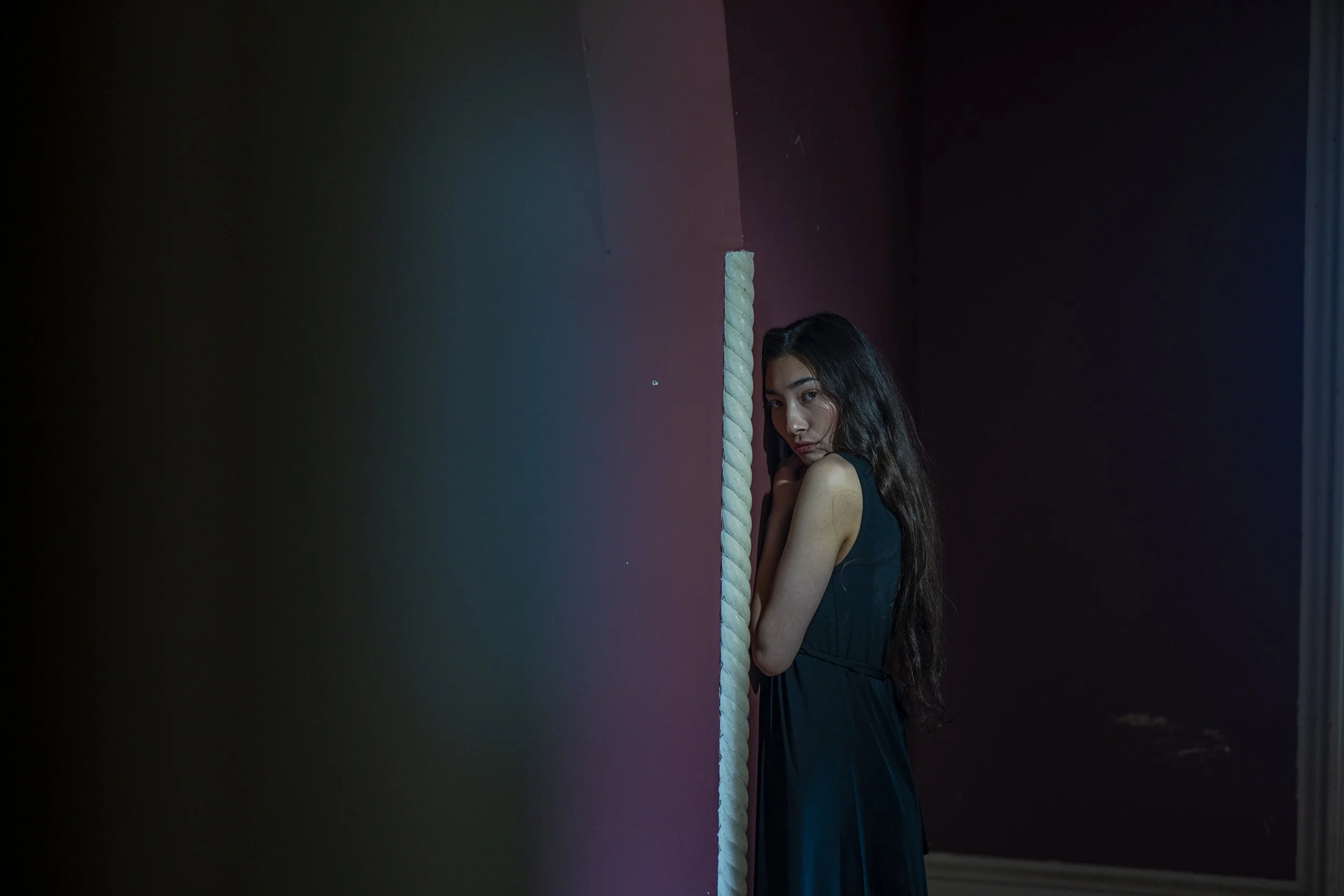 A young woman with long dark hair leaning against a wall, peering around a corner, in a dimly lit room with purple and dark blue walls.
