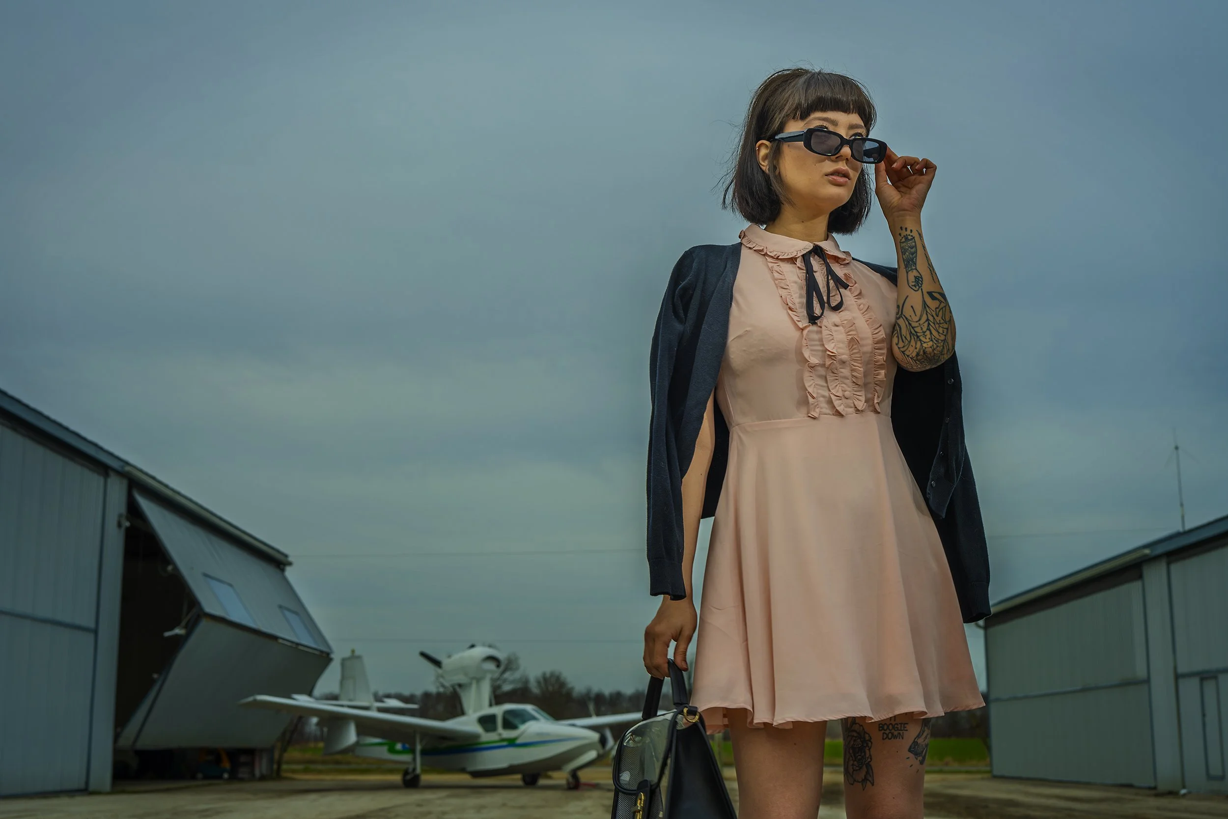 A woman in a pink dress and black jacket adjusting her sunglasses at an airport with small airplane and hangars in the background.