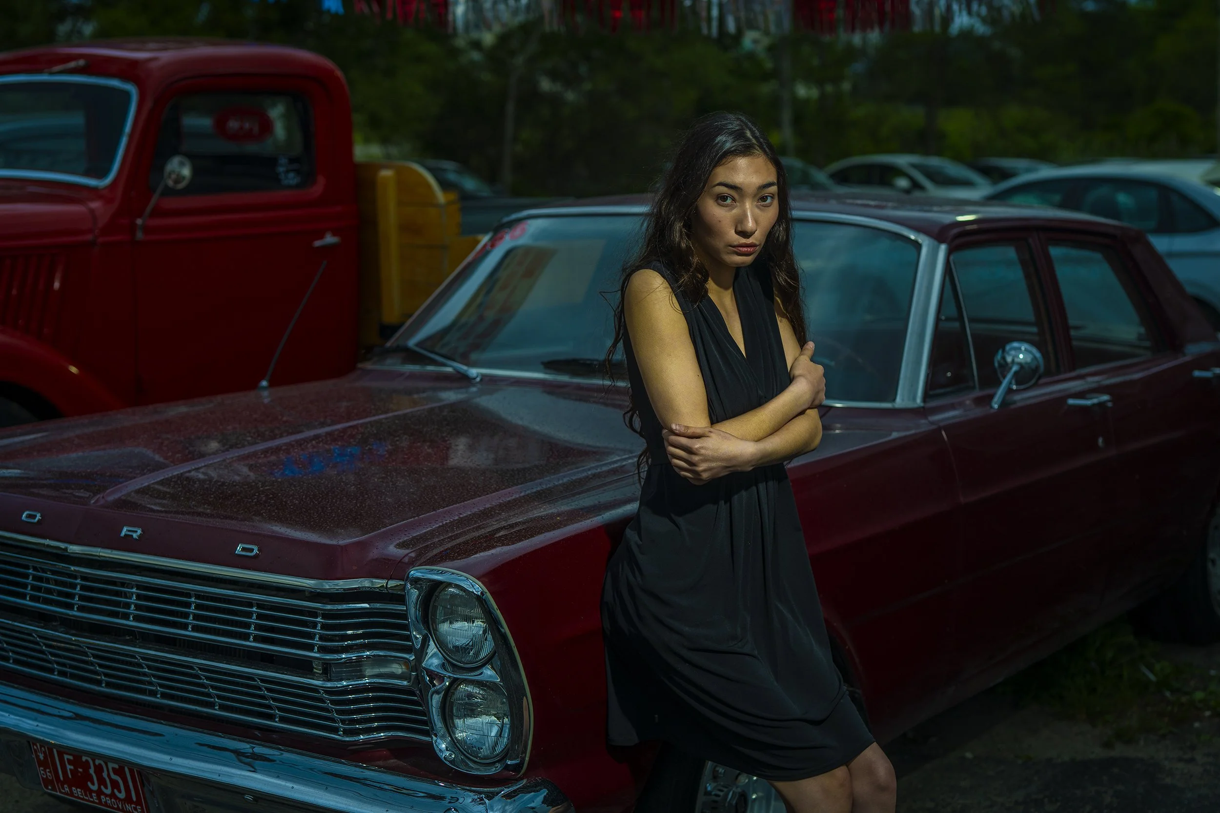 A woman standing next to a vintage red car with a dark exterior, arms crossed, looking at the camera with a serious expression in an outdoor setting with cars parked behind her.
