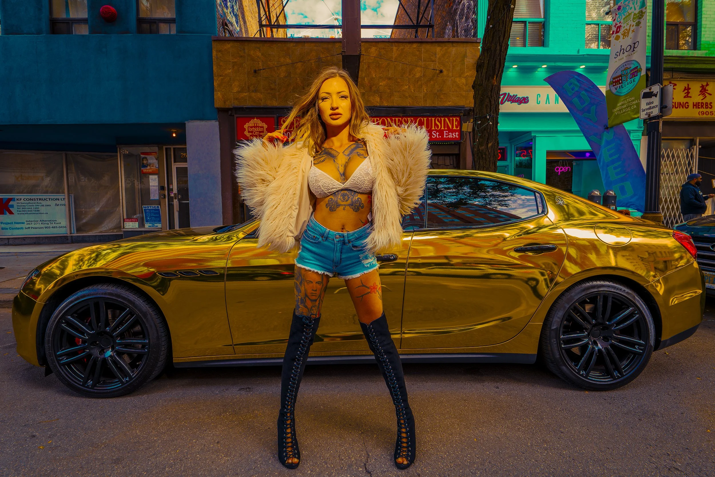 A woman with long blonde hair and tattoos on her chest and legs stands in front of a shiny gold car on a city street during daytime. She wears a white lace bra, distressed denim shorts, a fluffy beige jacket, and black thigh-high boots. The backgroun