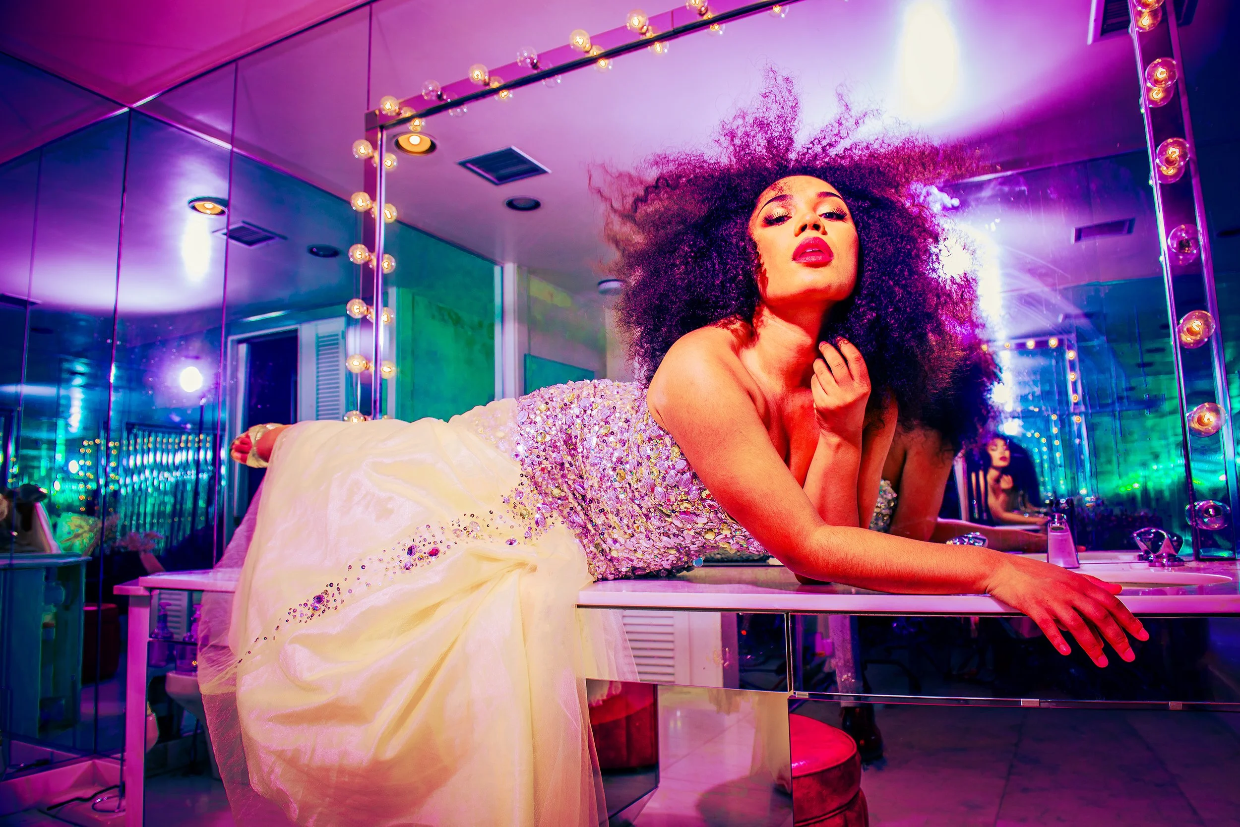 A woman with a large curly afro hairstyle wearing a shiny, beaded dress, lying on a white bed in front of a large mirror with light bulbs around it. The room is lit with colorful purple, pink, and green lights, creating a vibrant, energetic atmospher