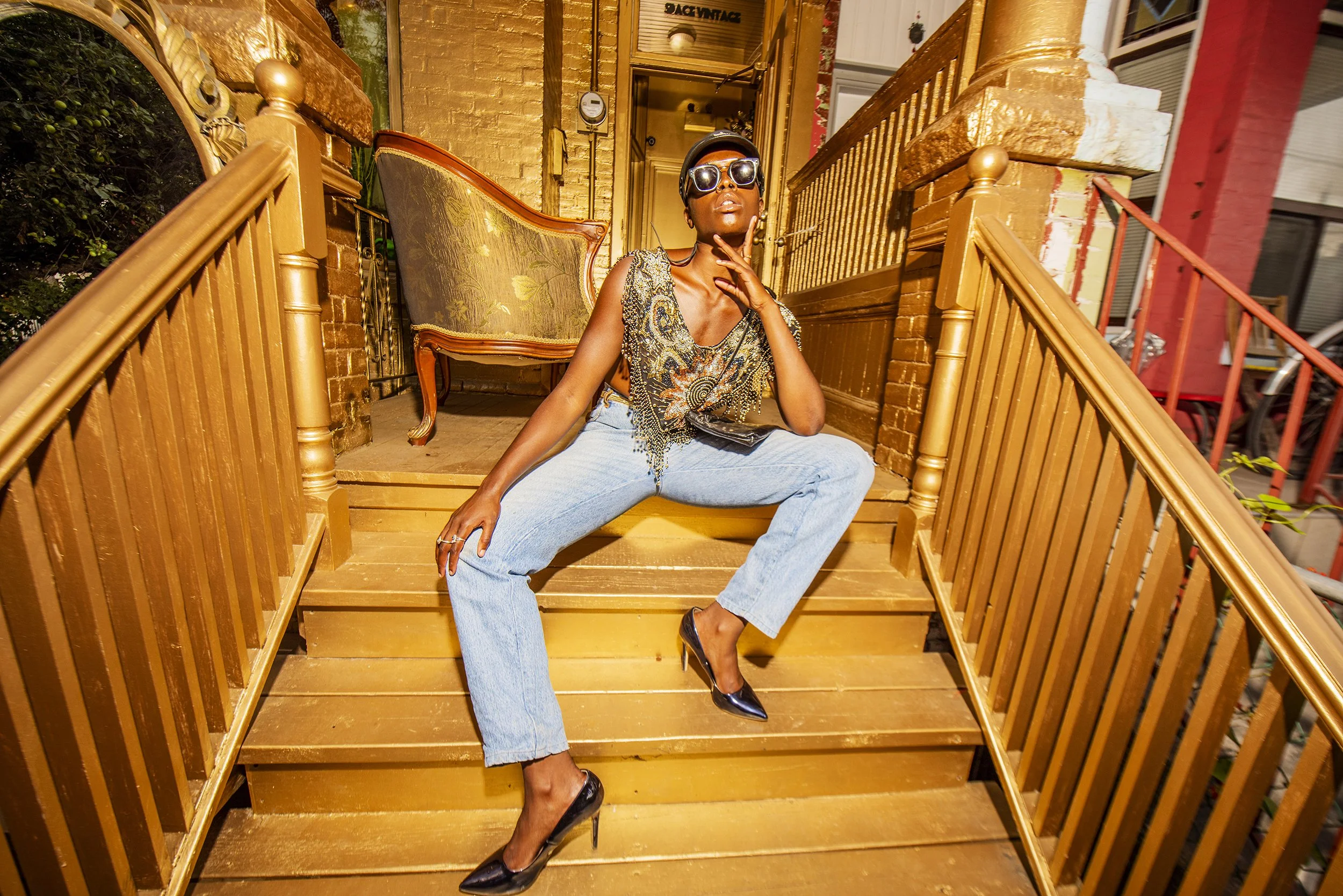 A woman dressed in a patterned top, light blue jeans, and black high heels, sitting on wooden stairs outside a house. She is wearing sunglasses, a hat, and jewelry, with a confident pose and hand near her face.