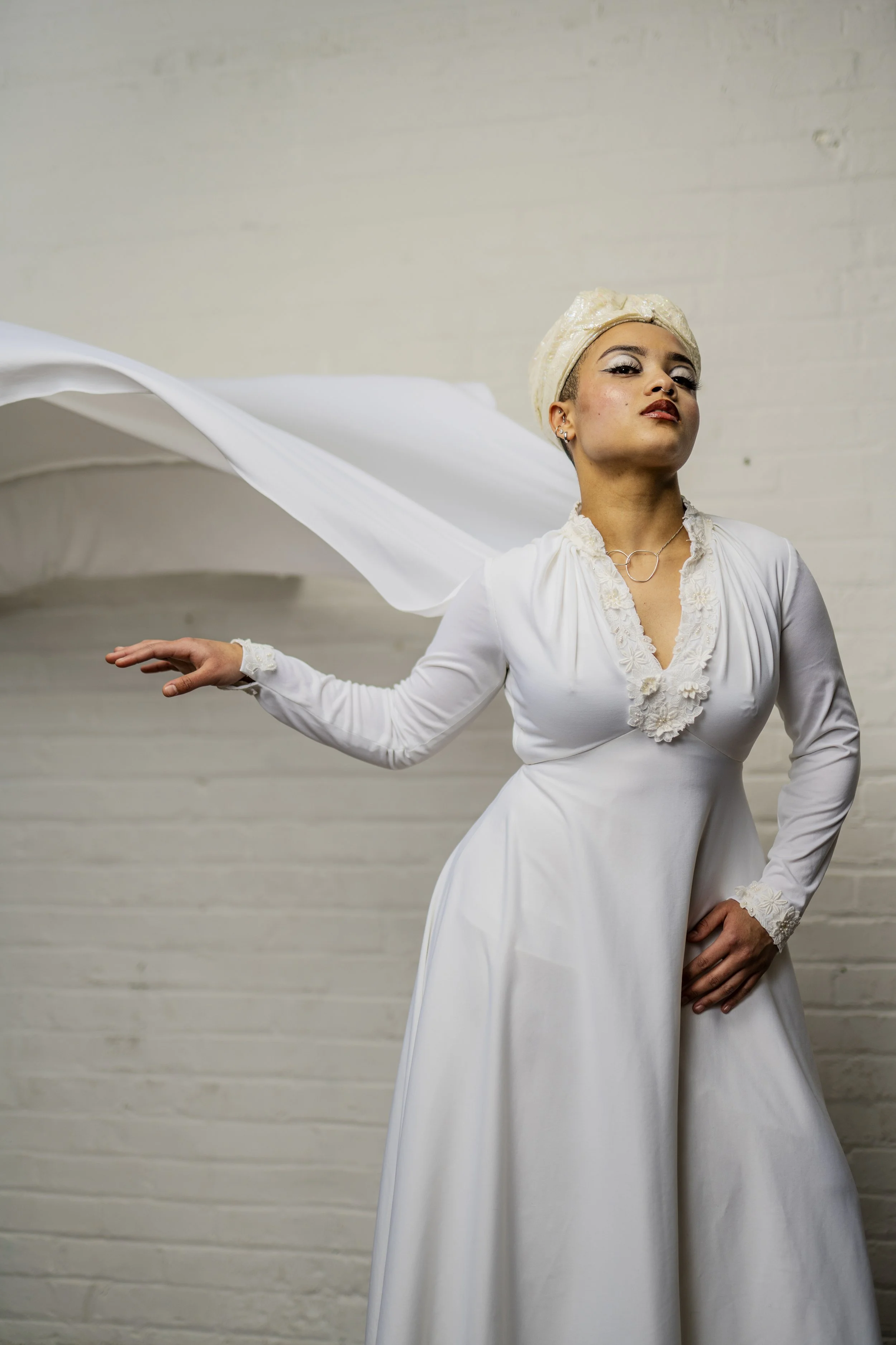A woman dressed in a white vintage-style dress with lace details and a headscarf, posing with her arm outstretched and hand open against a plain white brick wall.