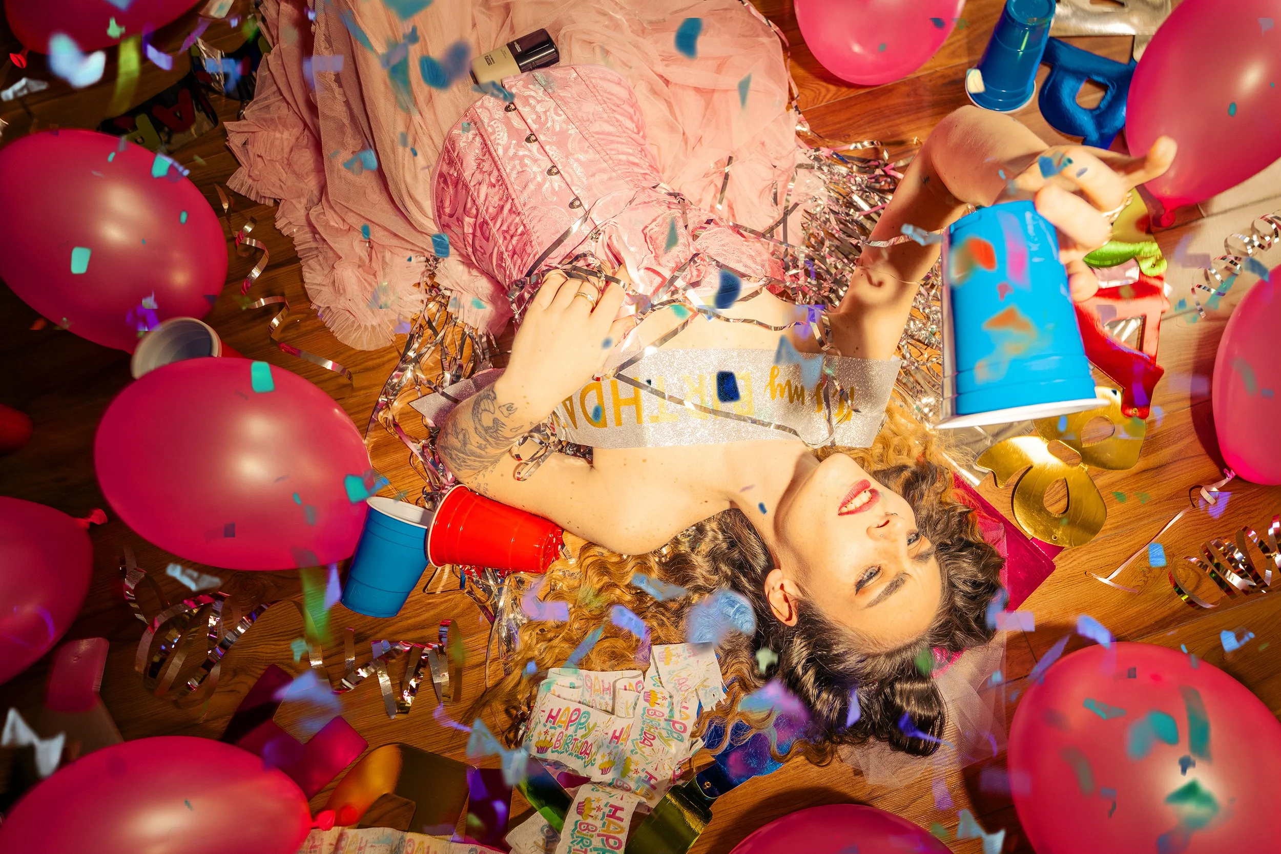 A woman with curly hair and tattoos lying on a wooden floor surrounded by pink balloons, confetti, party hats, and birthday decorations, celebrating a birthday party with a joyful expression.
