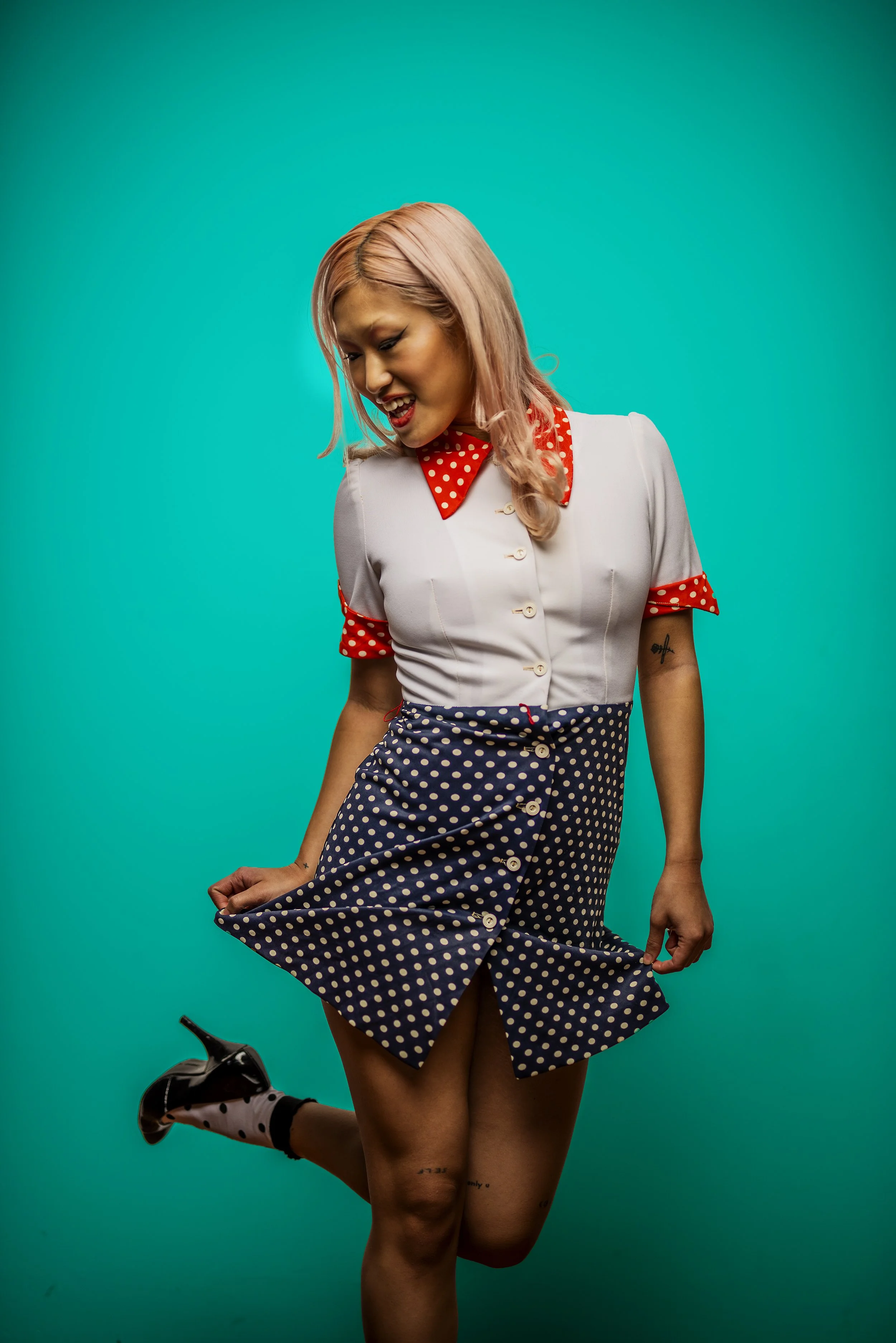 A woman with light pink hair smiling and looking down while lifting her polka dot skirt, dressed in a retro style outfit with a red polka dot collar and sleeves, standing against a teal background.