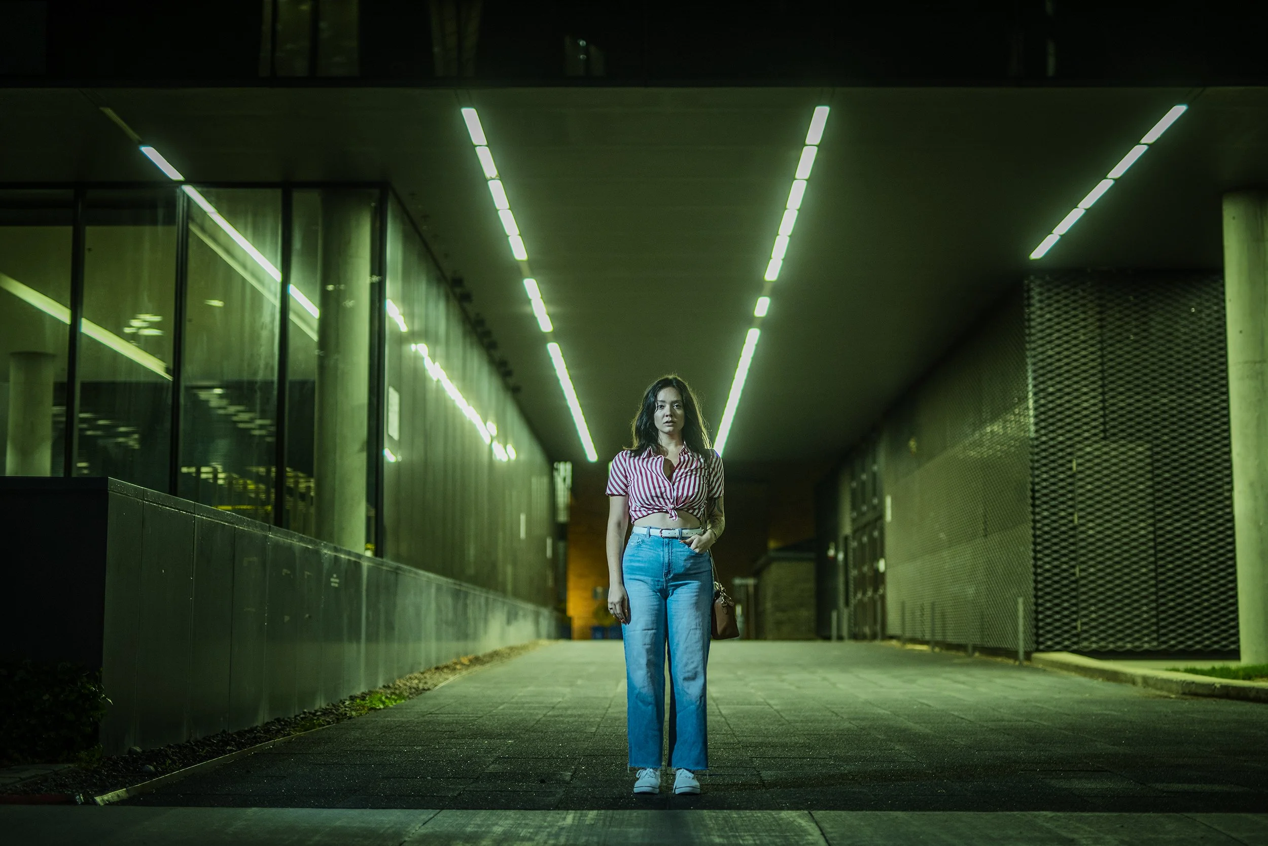A woman standing alone under modern green-lit architecture at night, wearing a striped red and white shirt, jeans, and white sneakers, holding a brown handbag.