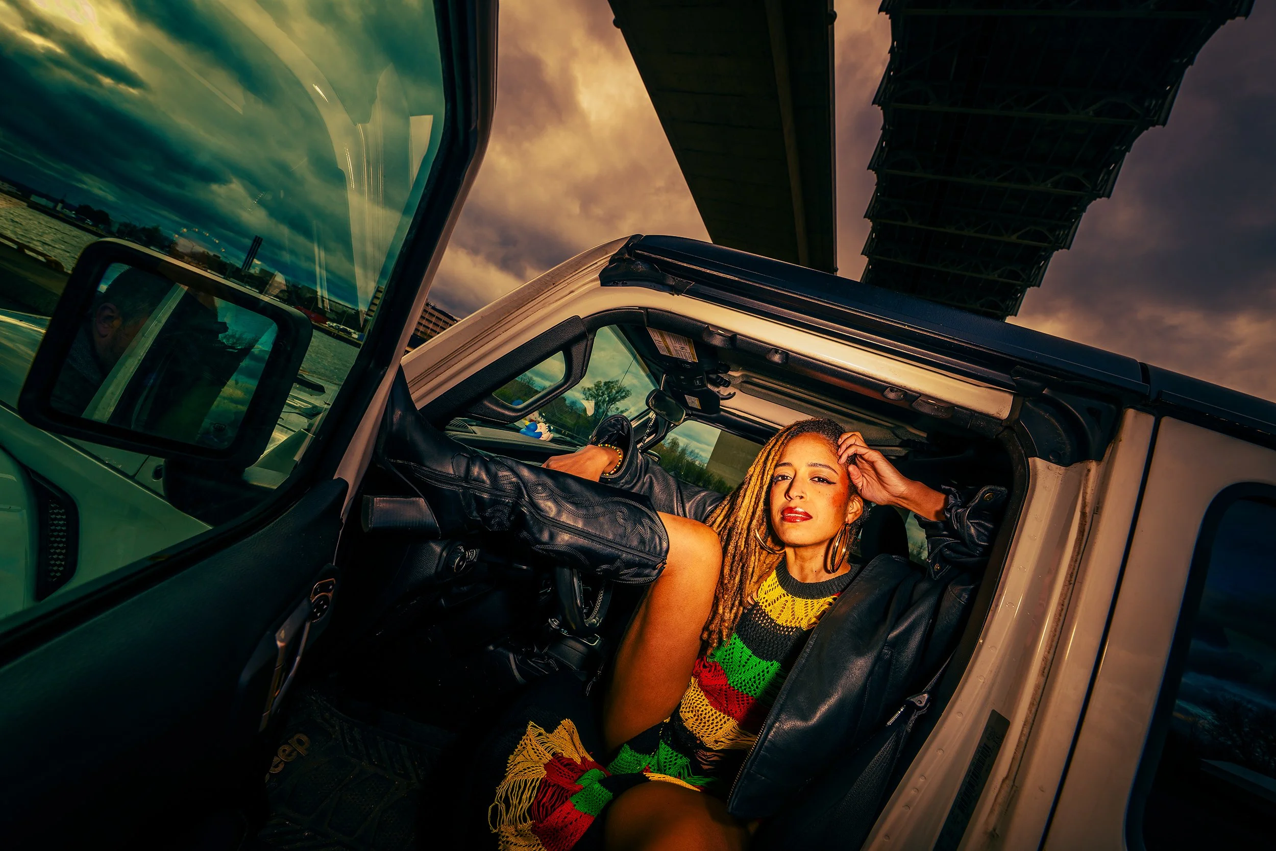 A woman with dyed blonde dreadlocks sitting in the driver’s seat of a Jeep, with her hand on her head, looking into the camera. The Jeep is parked under a bridge with a dramatic cloudy sky above.
