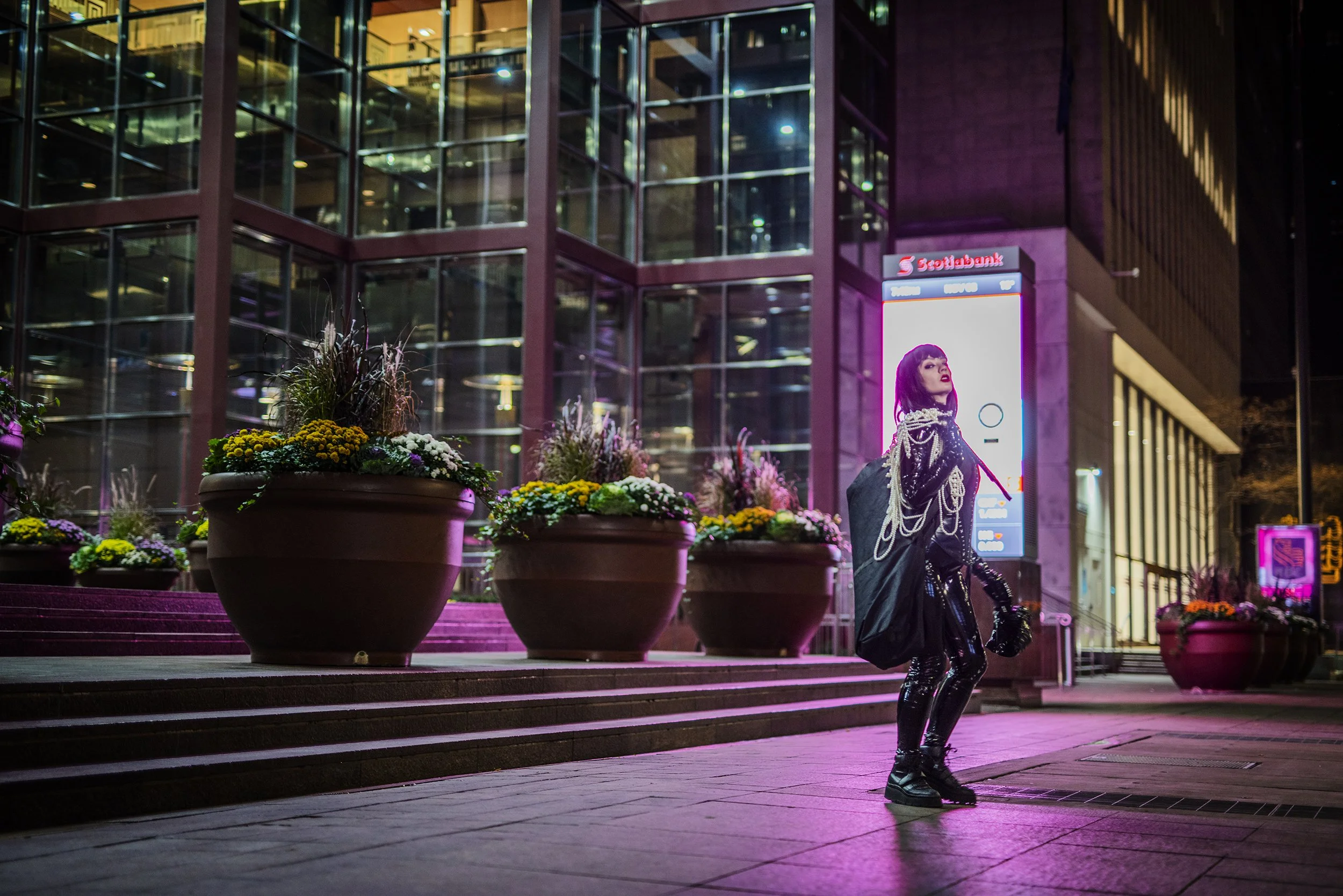 A person dressed in shiny black clothing and holding a large black bag stands on steps outside a modern building with glass windows and a digital billboard at night.