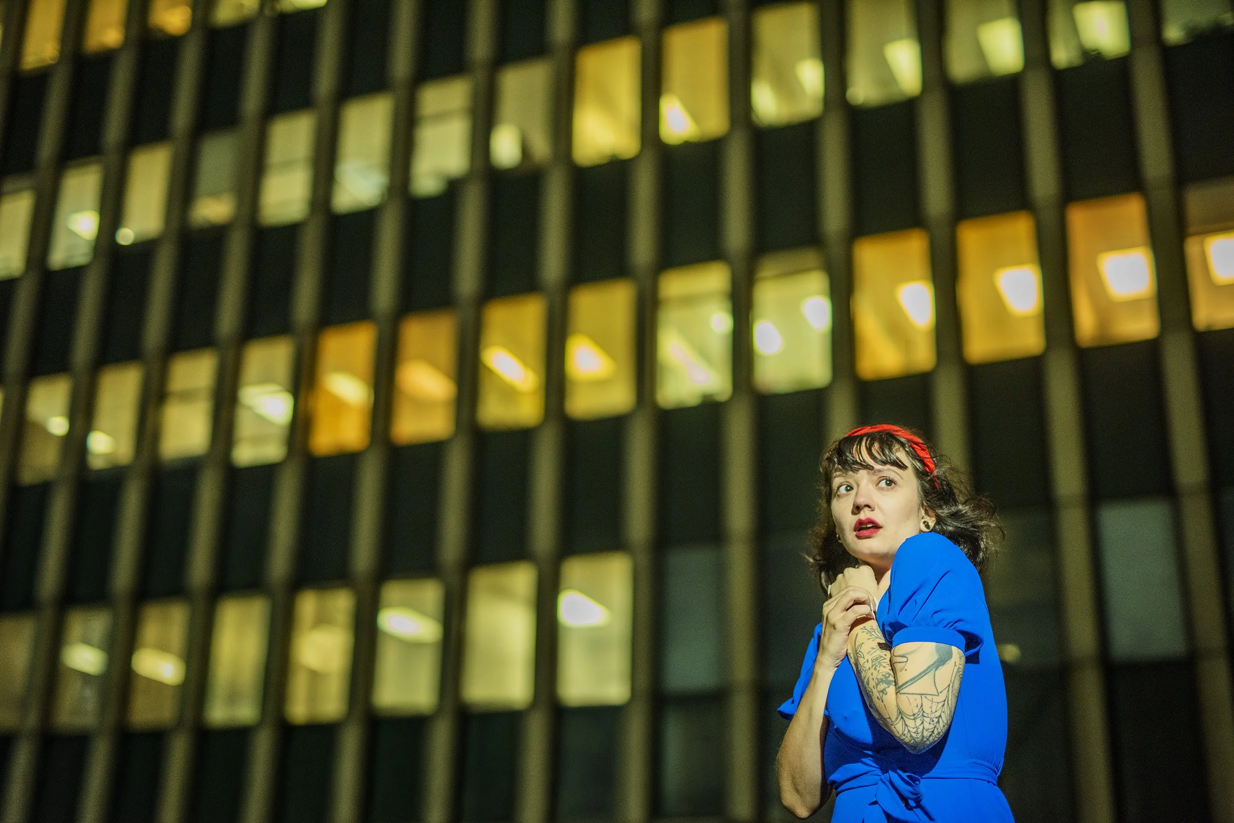 A woman with dark hair and tattoos on her arms wearing a blue dress, standing in front of a tall building with illuminated windows, looking off to the side with a concerned or surprised expression.