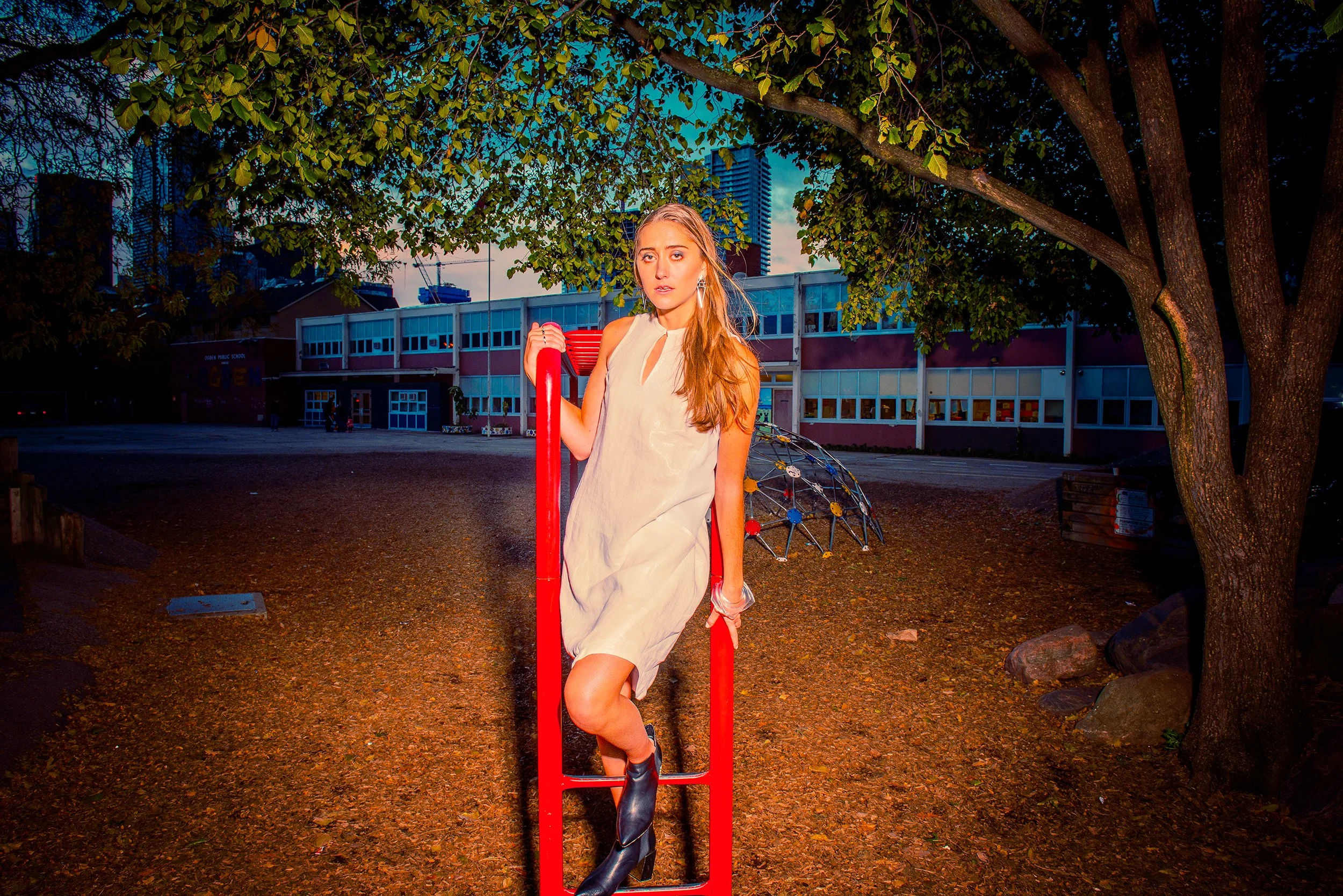 Young woman with long hair, wearing a beige dress and black boots, standing on a red playground structure under a large tree in a schoolyard at dusk.