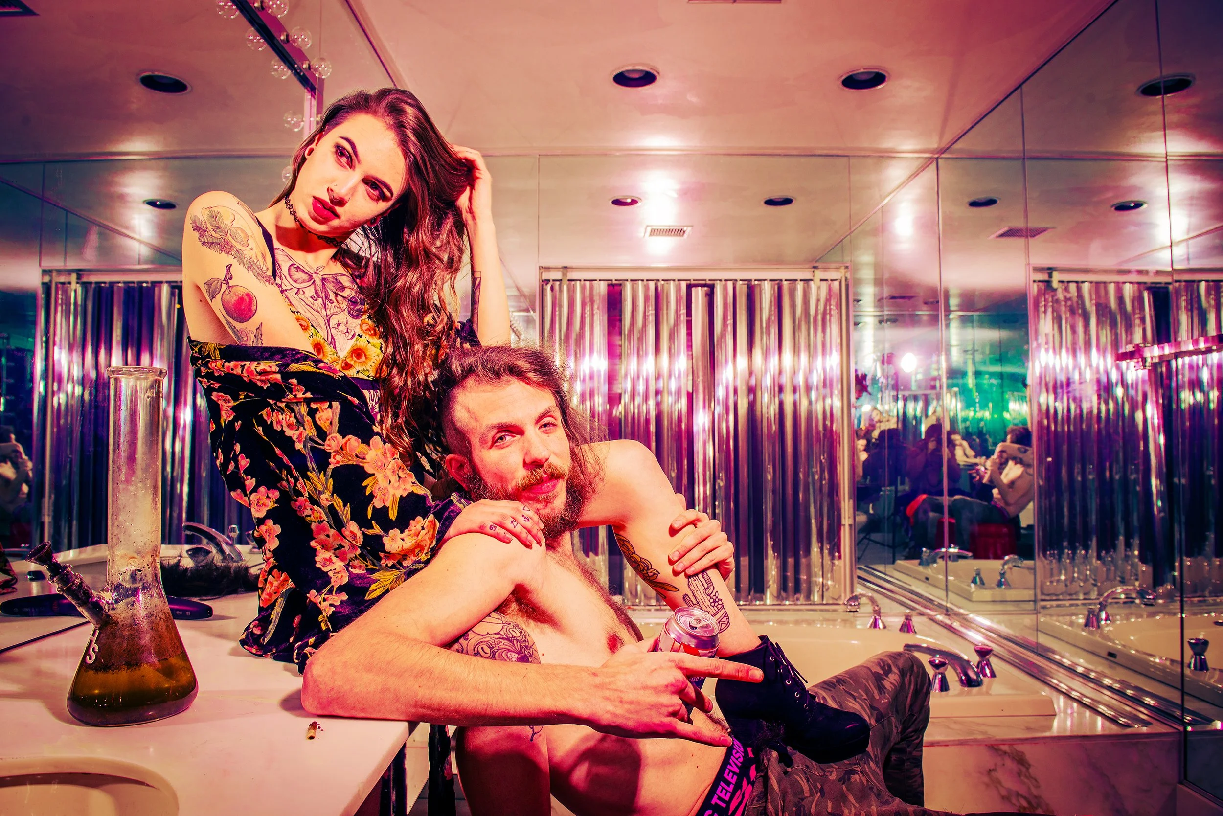 A woman with tattoos and dark hair, wearing a floral shirt, is sitting on a man's lap in a bathroom, with the woman in a playful pose and the man smiling. The bathroom has pink and purple lighting, mirrors on the walls, and a beaker and hookah on the