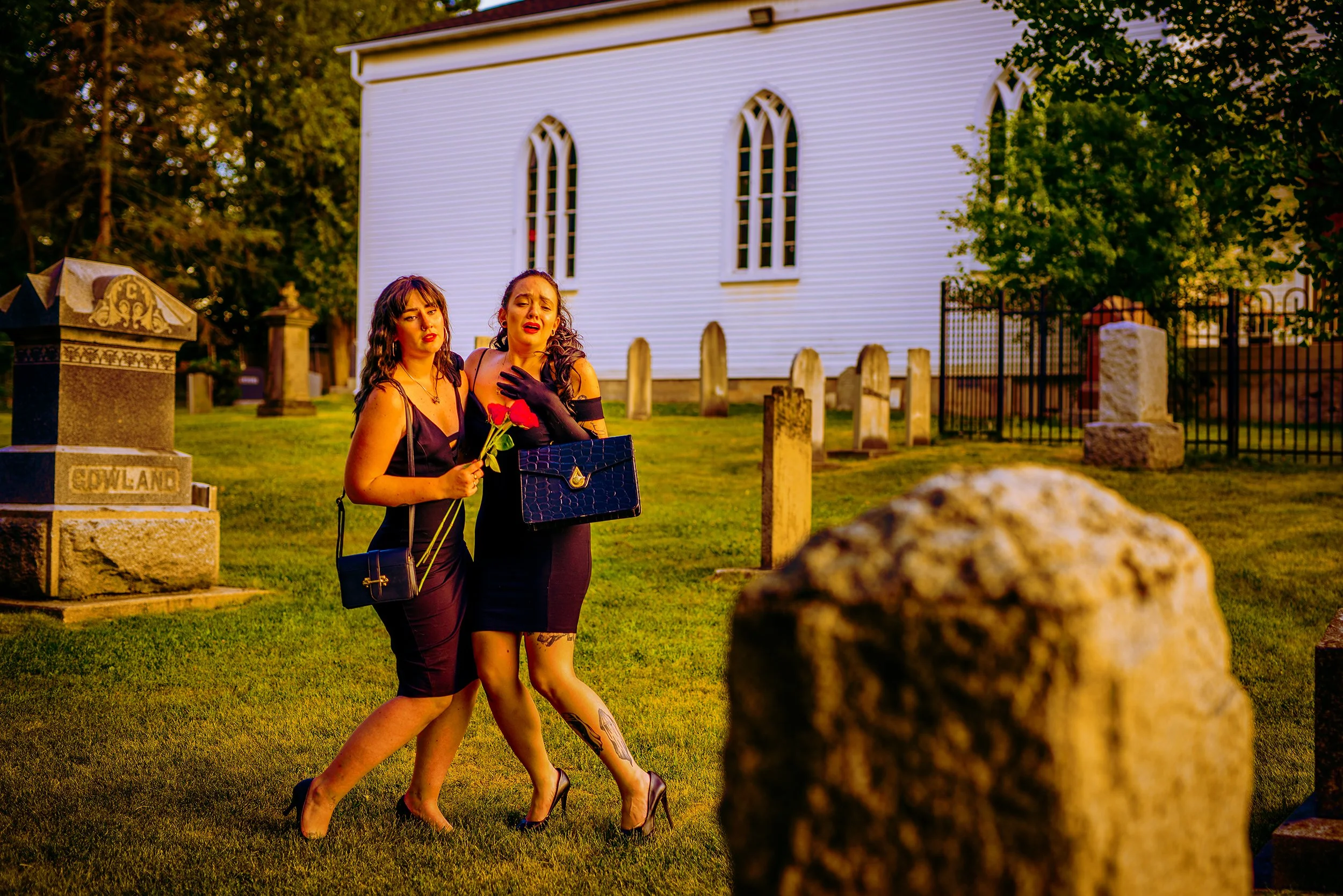 Two women dressed in black, holding red roses and carrying handbags, standing together in a cemetery during sunset, with a white church and gravestones in the background.