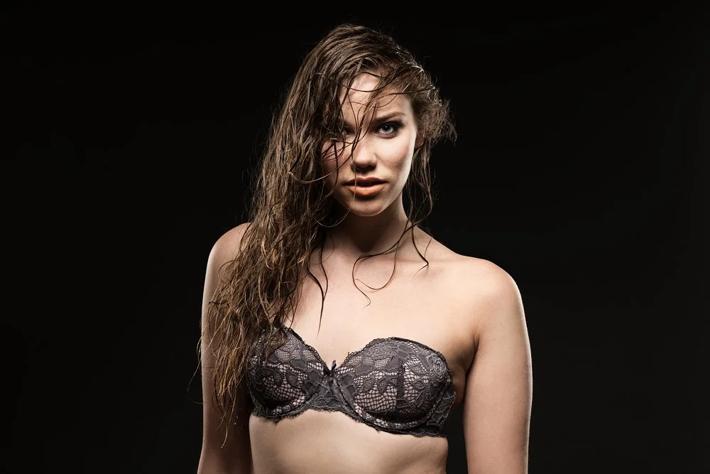 A woman with wet, curly hair wearing a strapless black lace bra, looking at the camera against a dark background.