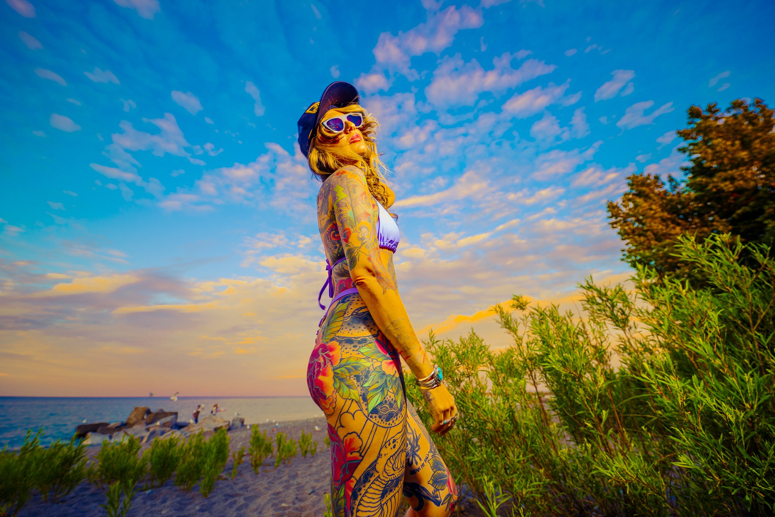 A woman with tattoos wearing a bikini, sunglasses, and a cap standing on beach during sunset, with sky, ocean, and greenery in the background.