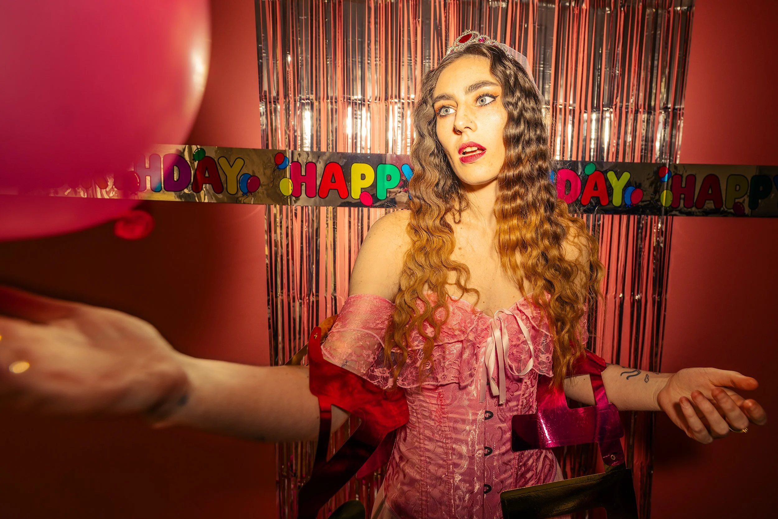 A woman with long curly red hair wearing a pink dress and a tiara at a birthday party, standing in front of a 'HAPPY BIRTHDAY' banner and metallic streamers, with one arm extended outward.