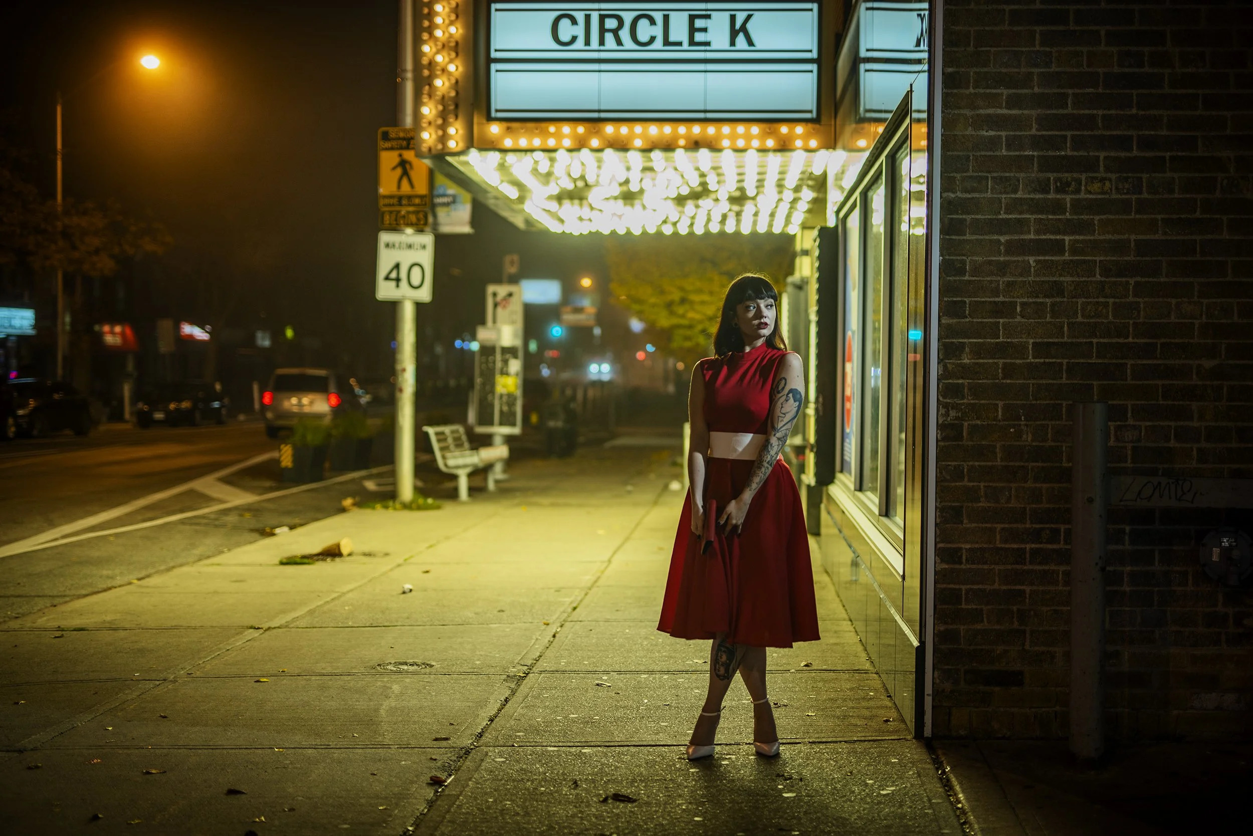 A woman in a red dress and heels standing outside a movie theater at night, illuminated by marquee lights, with a brick wall on one side and cars in the background.