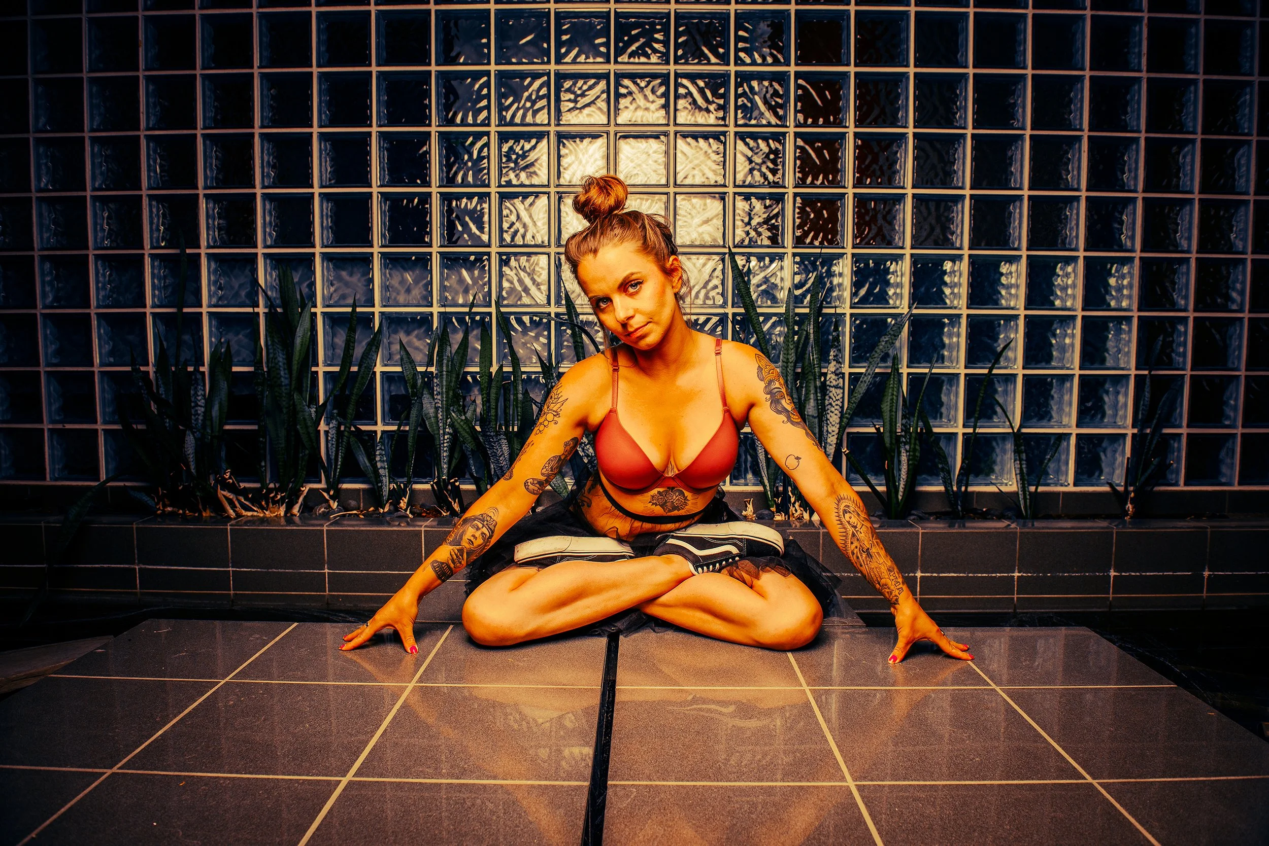 A young woman with tattoos on her arms and chest, sitting on the ground with crossed legs, leaning on her arms, in front of a glass brick wall with plants behind her, wearing a red top and black shorts.