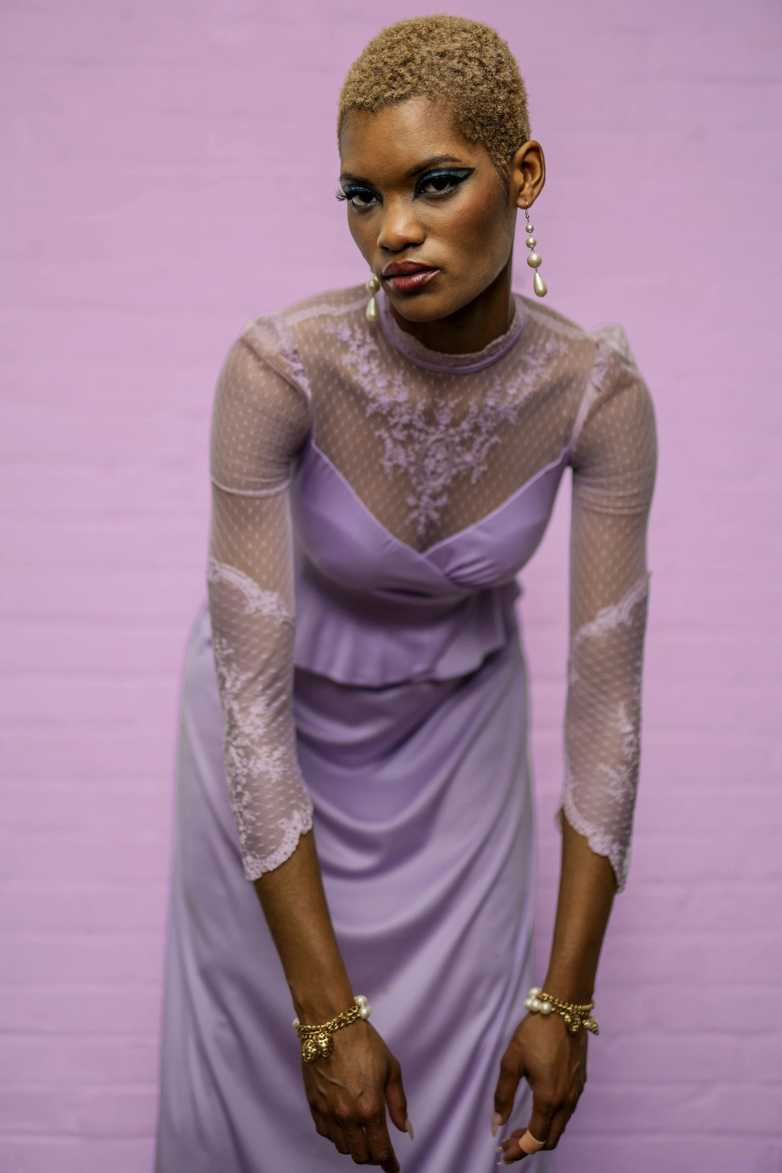 A woman with short, blonde hair and bold makeup, wearing a lavender dress with sheer lace sleeves, and pearl earrings, is posing against a pink background.